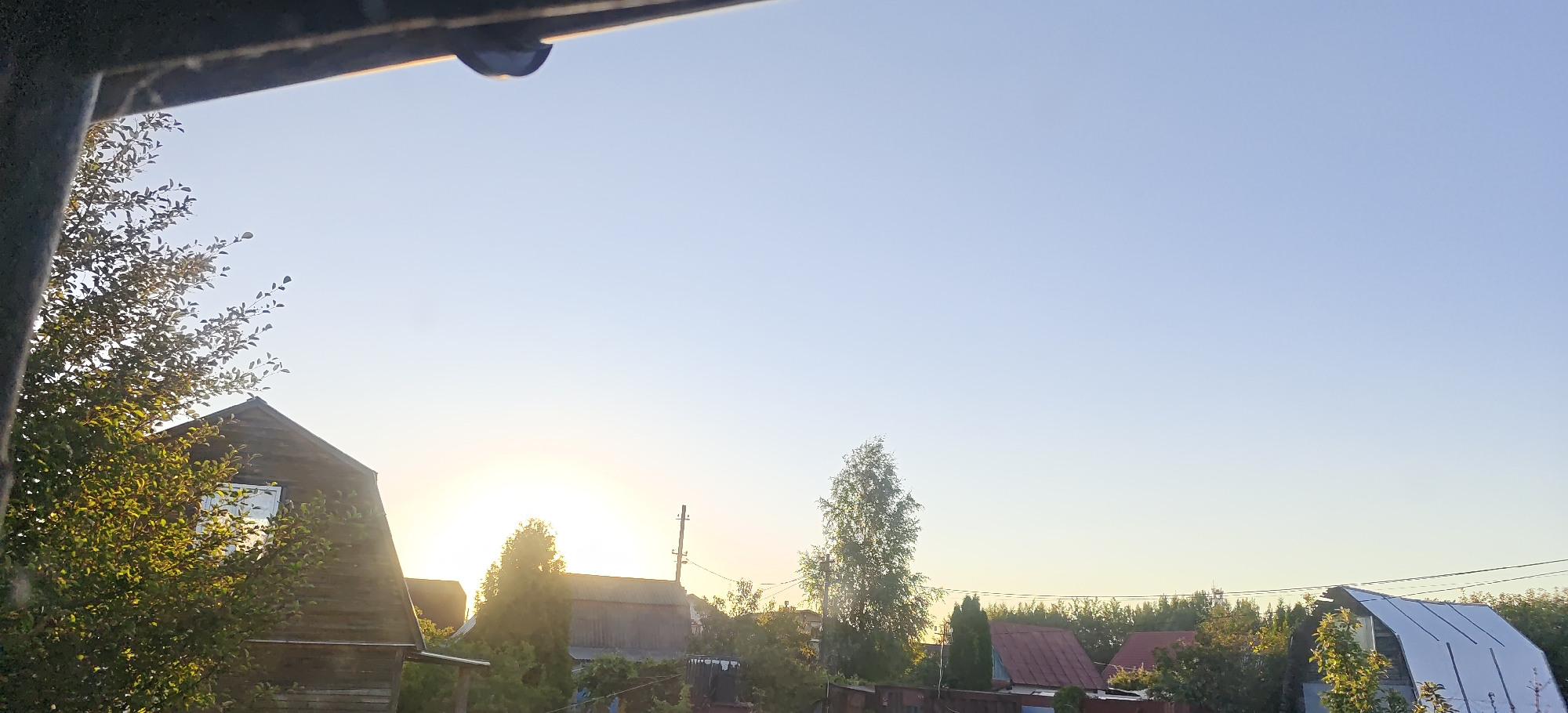 The image is a wide outdoor shot of a rural village at sunset. In the foreground, a dark, angled structure, possibly a roof or overhang, partially obscures the top of the frame. To the left, a wooden house with a sloped roof and a visible window is partially covered by a dense green tree with many leaves. The setting sun is visible in the distance, behind a line of trees and houses, casting a warm glow. Several other houses with various roof colors and materials are scattered throughout the middle ground, interspersed with trees and foliage. A power line runs horizontally across the middle of the frame, and a single utility pole stands out on the right. The sky is a clear, pale blue, transitioning to a lighter hue near the horizon. The overall scene depicts a serene, twilight atmosphere in a tranquil village setting.