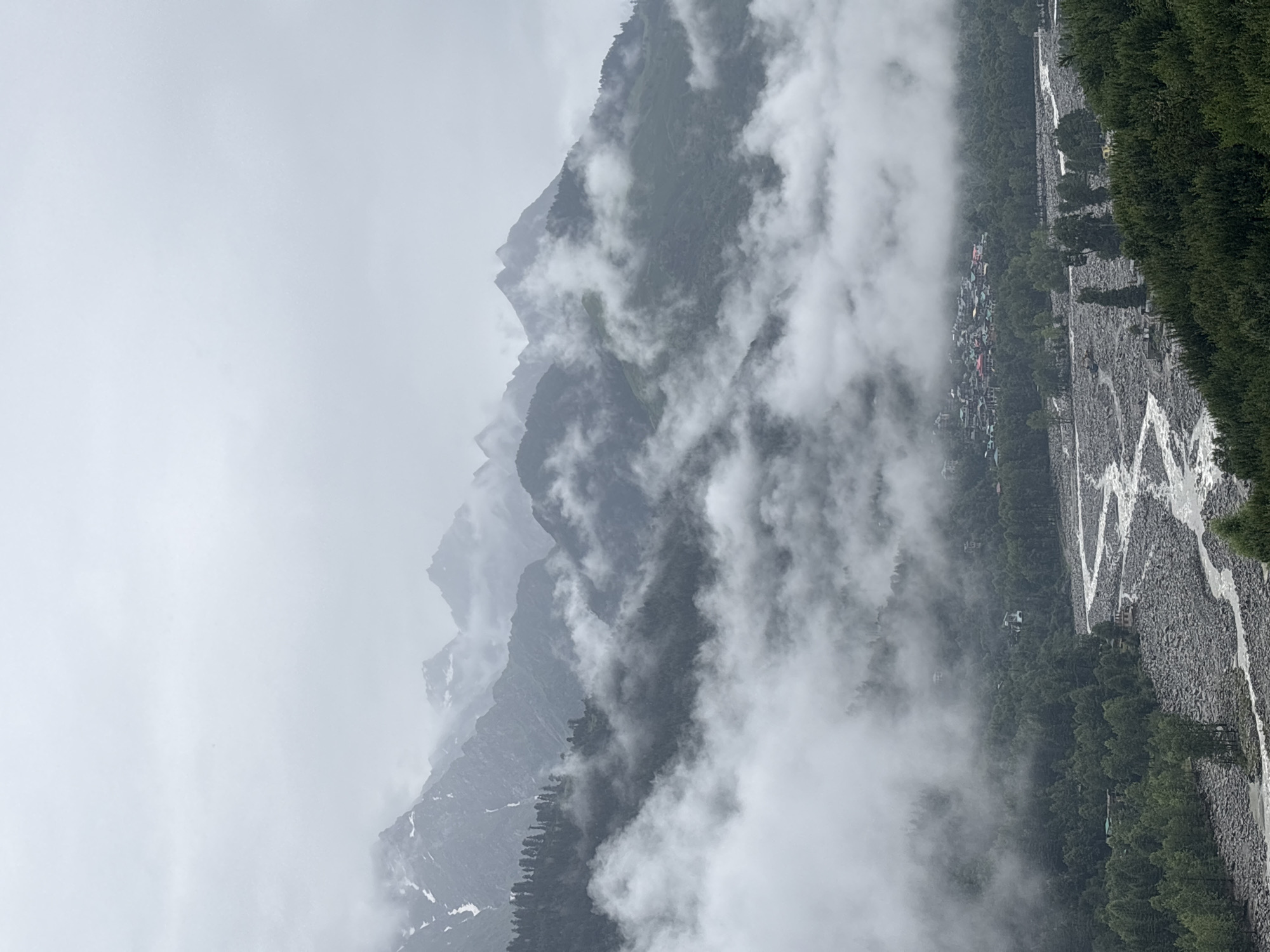 The image depicts a cloudy and foggy mountain landscape with a river flowing through a valley. In the distance, a small town, identified as Vashisht, India, is visible nestled among the trees. The town features buildings with red and green roofs. The river is wide and appears to be flowing over a rocky bed, with some trees lining its banks and extending up the surrounding hillsides. The mountains in the background are partially obscured by clouds and fog, with patches of snow visible on some of the peaks. The overall scene conveys a sense of natural beauty and tranquility.