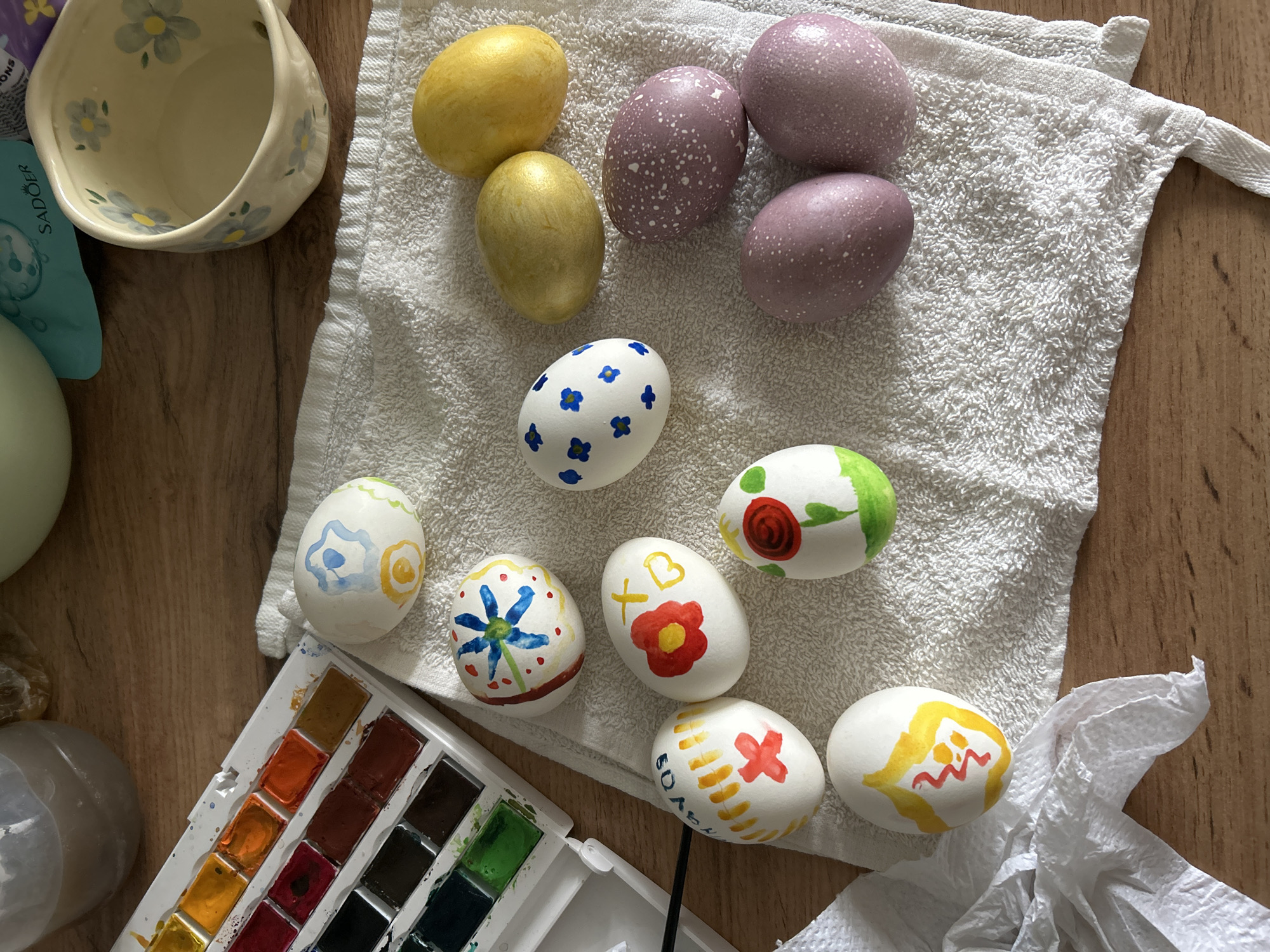 The image shows an array of decorated Easter eggs resting on a white towel on a wooden surface. There are two yellow eggs, three mauve eggs with white specks, and several white eggs decorated with painted designs in blue, red, yellow, and green. A small floral-patterned bowl is visible in the upper left corner, and a watercolor paint palette is in the lower left. A small portion of a white stone-like object can be seen in the lower left corner. The image was captured in Chaikovsky, Russia.