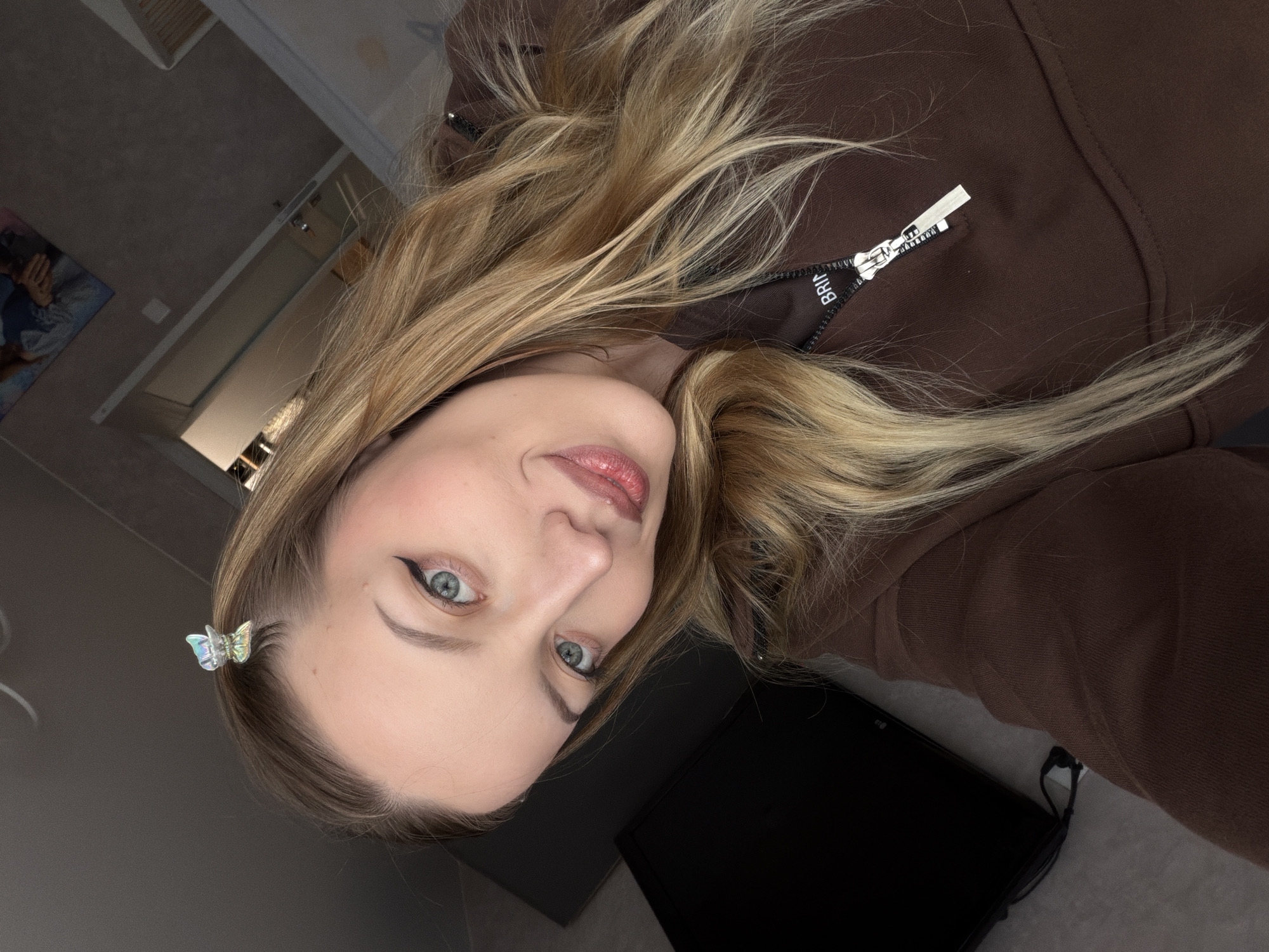 A woman is positioned at an unusual angle in the frame, her face tilted upwards towards the camera. She has fair skin, light blonde hair with highlights, and is wearing black eyeliner. A butterfly clip adorns her hair near her temple. She is wearing a brown hooded sweatshirt. Behind her is a dark object, likely a monitor or laptop, and a glimpse of a room with neutral walls and what appears to be a painting or picture in the background. There is no indication that the photo was taken in the city of Reshetnikovo, Russia.