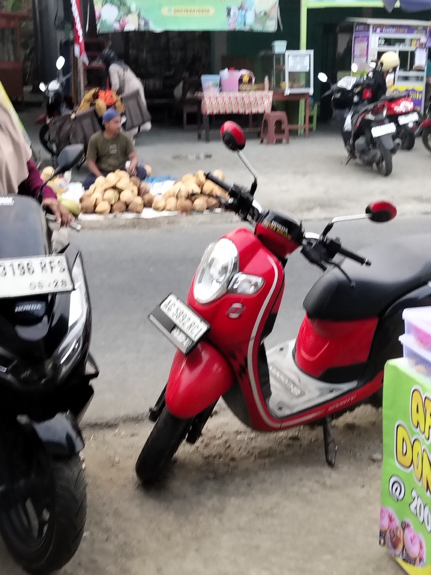 A street scene in Gempolan, Indonesia, features a vendor selling coconuts from a roadside stall. In the foreground, a person in a maroon hijab and a purple shirt is partially visible, with their hand resting on a coconut. Behind them, a man wearing a green t-shirt and a blue cap sits cross-legged amidst a large pile of coconuts. Two motorcycles are parked near the stall; a black one on the left with a license plate reading "B 136 KFS" and a red one on the right with a license plate reading "AG 3892 RCI." In the background, several other motorcycles are parked, and a woman in a light-colored headscarf and clothing is attending to a stall. A green cart with a sign advertising "APEL DONUT" with a price of "@ 200" is visible to the right of the red motorcycle. The ground is paved with concrete, and there are small buildings and awnings in the background.