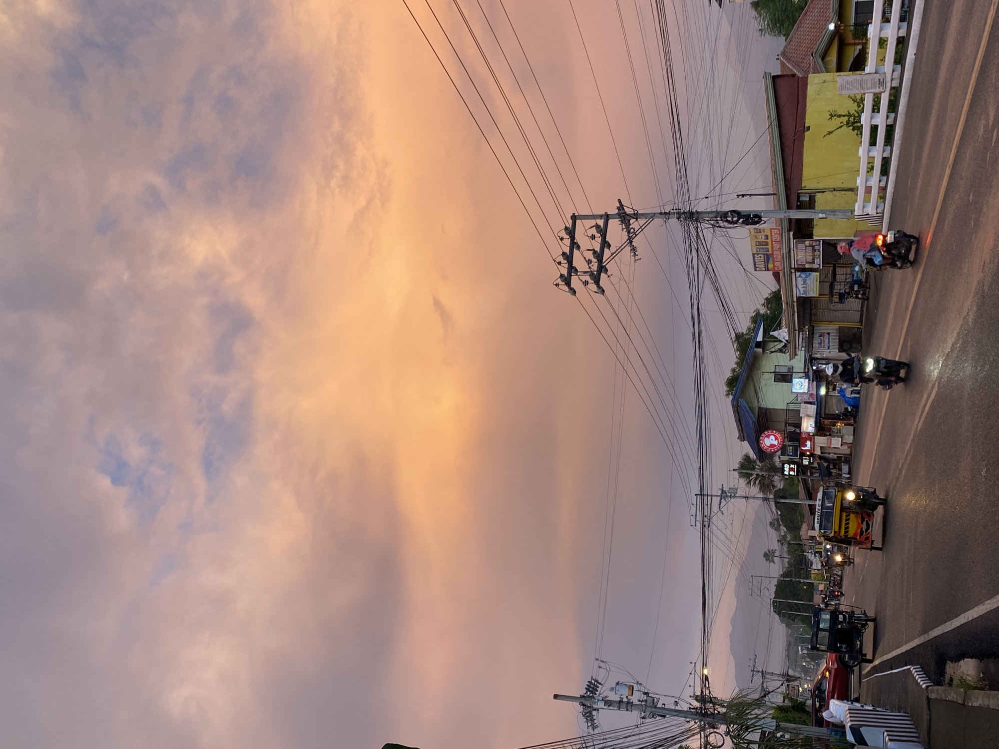 The image depicts a street scene in Davao City, Philippines, during sunset. The sky is filled with clouds tinged with hues of orange, pink, and purple. The road is paved and wet, reflecting the warm colors of the sky.
On the road, several vehicles are visible. Two tricycles, one yellow and one black, are moving forward. A motorcycle with a rider wearing a helmet is on the right side of the road, also heading forward. Another motorcycle is seen in the distance.
Buildings line the street, featuring a mix of commercial establishments. One building has a yellow facade with a red roof, and another has a green and yellow exterior. Signs on the buildings include "DAVIES PAINT CENTER" and a circular red logo with white text. Utility poles with numerous wires are prominent along the street.
In the background, mountains are faintly visible, shrouded in the twilight haze. Palm trees and other vegetation are scattered throughout the scene. The overall atmosphere suggests a busy street in a tropical urban environment at dusk.