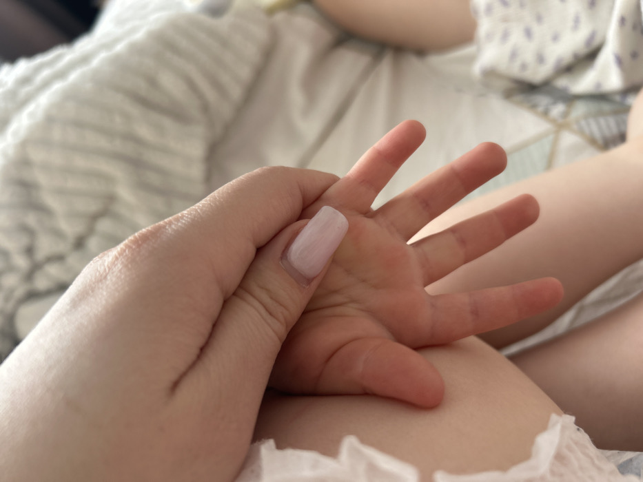 The image captures a close-up view of an adult's hand interacting with an infant's hand and leg. The adult's light-skinned hand, positioned on the left and center-top, features a single fingernail with a square shape, painted in a light pink or nude shade, suggesting it is either an acrylic or gel extension. This hand appears to be gently resting against or holding the infant's small, light-skinned hand, which is slightly splayed in the center of the frame. To the left, a portion of the infant's leg or thigh is visible, along with the white, ruffled edge of what appears to be a diaper or garment. The background consists of soft-focus elements: a white, textured fabric (possibly a blanket or bedding) in the upper right, and a patterned fabric with light abstract shapes (possibly a blanket or clothing) in the lower right. The scene is indoors, likely a domestic setting such as a bedroom. The specific location within the city of Chaikovsky, Russia, cannot be determined from the visual information presented in the image.