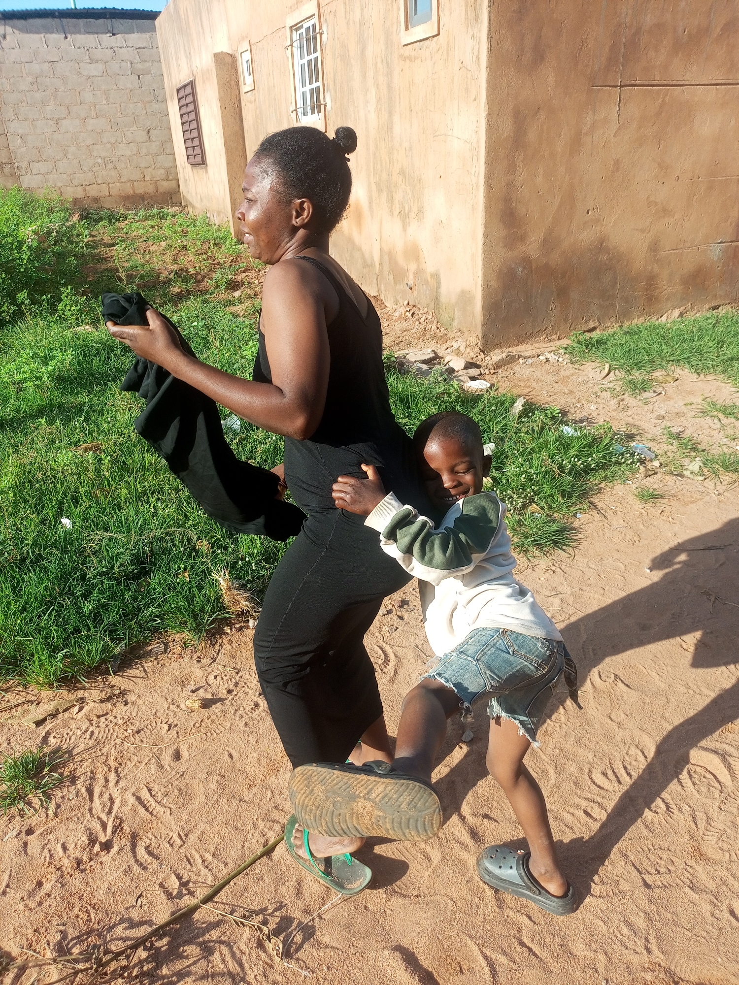 In a sandy, outdoor area with patchy green grass and a tan building in the background, a woman wearing a black sleeveless dress and black pants is holding a black garment in her left hand. A young child, wearing a white and green long-sleeved shirt and ripped denim shorts, is hugging the woman from behind and resting their head on her back. The child is barefoot. The woman is looking to her left. The scene appears to be in Tempure, Nigeria.