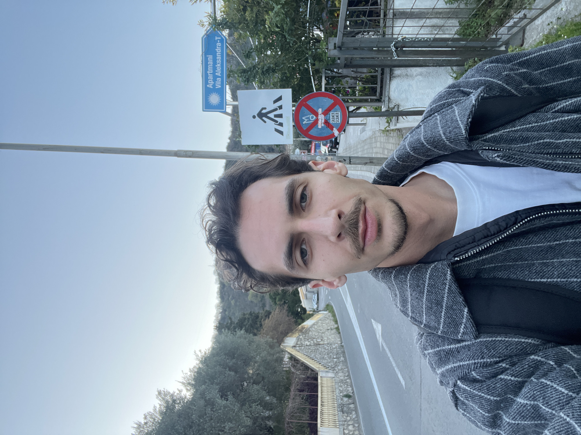 A man takes a selfie in Budva, Montenegro. He stands on a roadside next to pedestrian and traffic signs, near a road with a white arrow marking. In the background are trees, a stone wall with a railing, and a glimpse of buildings and sky. The signs read "Apartmani Vila Aleksandra-T", depicting a sun, and have a pedestrian crossing symbol, and a circular sign depicting a man operating a vehicle with a red line through it, possibly indicating a prohibition of certain vehicles. The man wears a white shirt and a gray and black striped jacket.