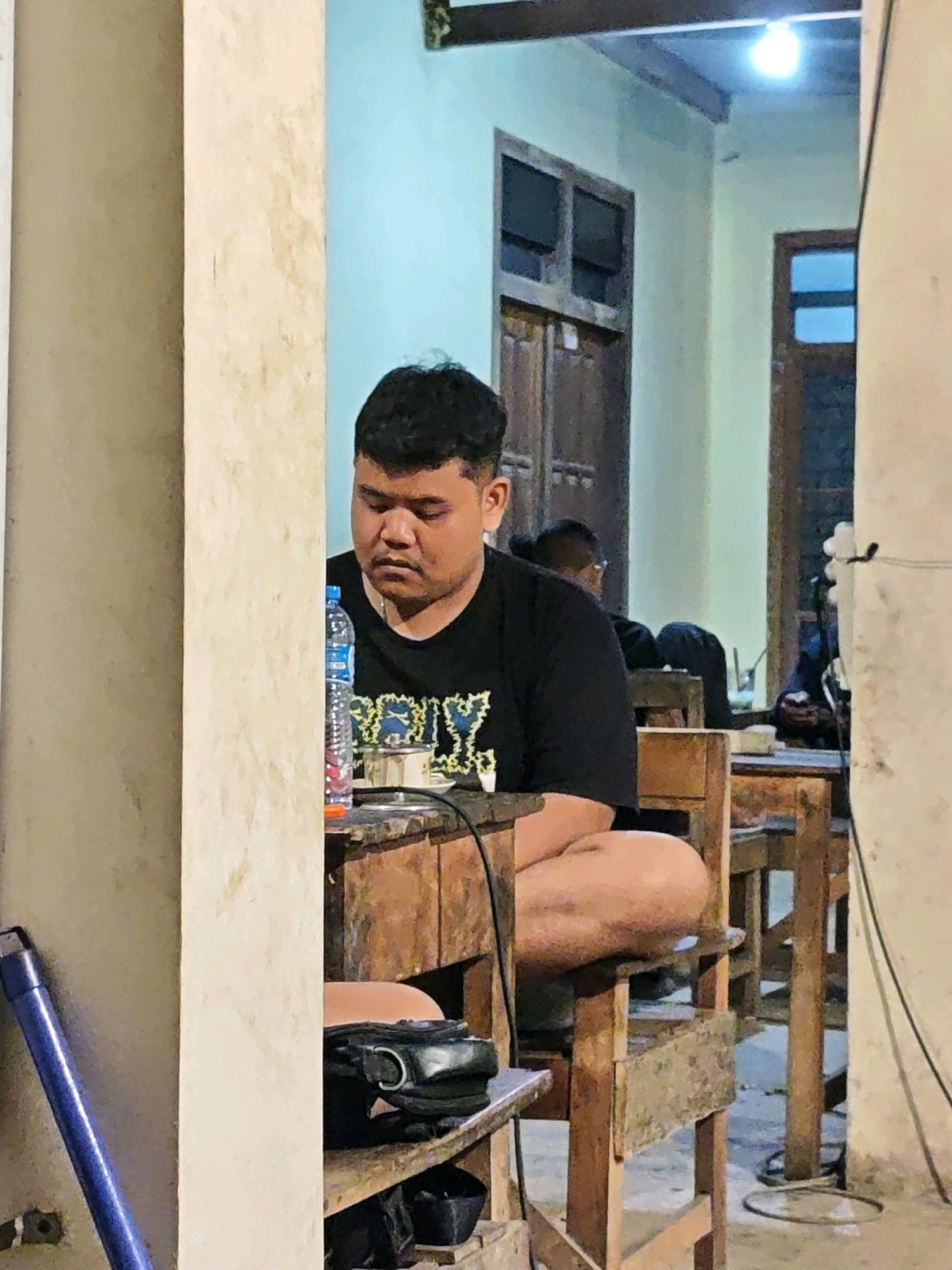 The image shows a man sitting at a wooden table, his head bowed. He is wearing a black t-shirt with a design on the front. A bottle of water and a small bowl are on the table in front of him. The background suggests an indoor setting with wooden furniture and doors. The overall atmosphere appears to be casual and possibly informal. The man is the primary subject, with the environment providing context.