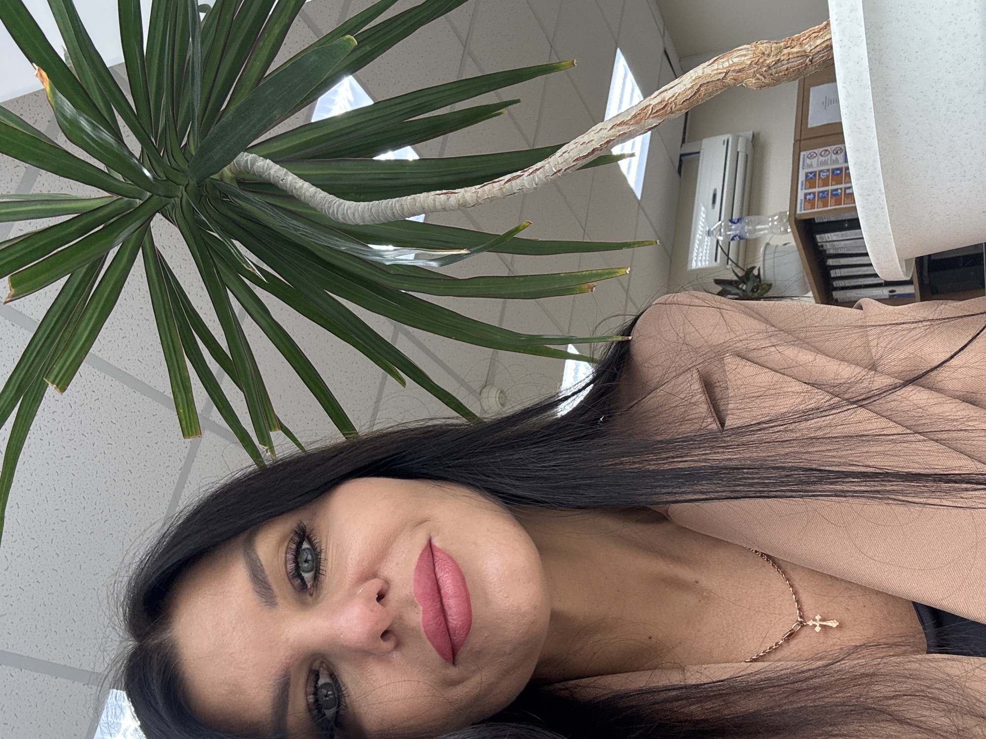 A woman with dark hair is posing for a selfie. She is wearing a beige top and a gold necklace with a cross pendant. A potted Dracaena plant is partially visible in the upper left corner of the frame. The background appears to be an office setting with a white ceiling, fluorescent lights, and a storage unit. The location is likely in an office building in the city of Chaikovsky, Russia.