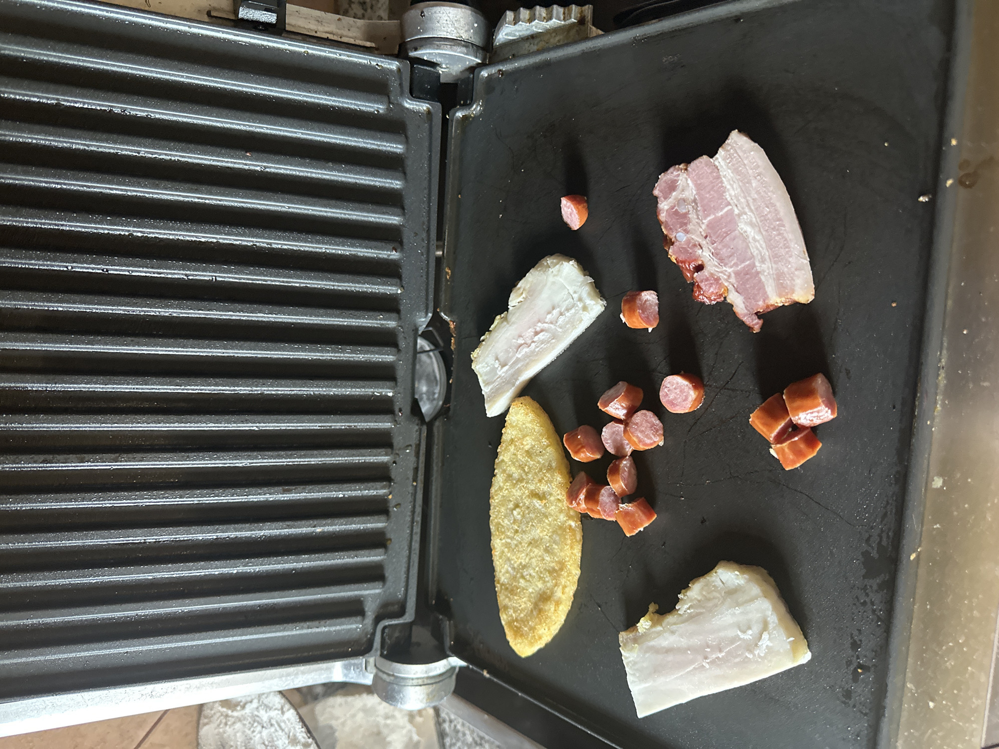 An opened electric grill contains food items on its lower plate. The grill plate is dark gray and ribbed. On the left side of the plate, there is a piece of white fat. Next to it, a cluster of sliced red sausages is visible. To the right of the sausages, there is a piece of raw bacon with visible layers of fat and meat. In the center of the plate, a thin, oval-shaped, golden-brown food item, possibly a hash brown or latke, lies flat. Behind the hash brown, another piece of white fat is positioned. A few more sliced sausages are scattered around the plate. The upper plate of the grill is raised, showing its ribbed surface. The background is blurred, suggesting an indoor setting with a neutral-colored wall and countertop.
