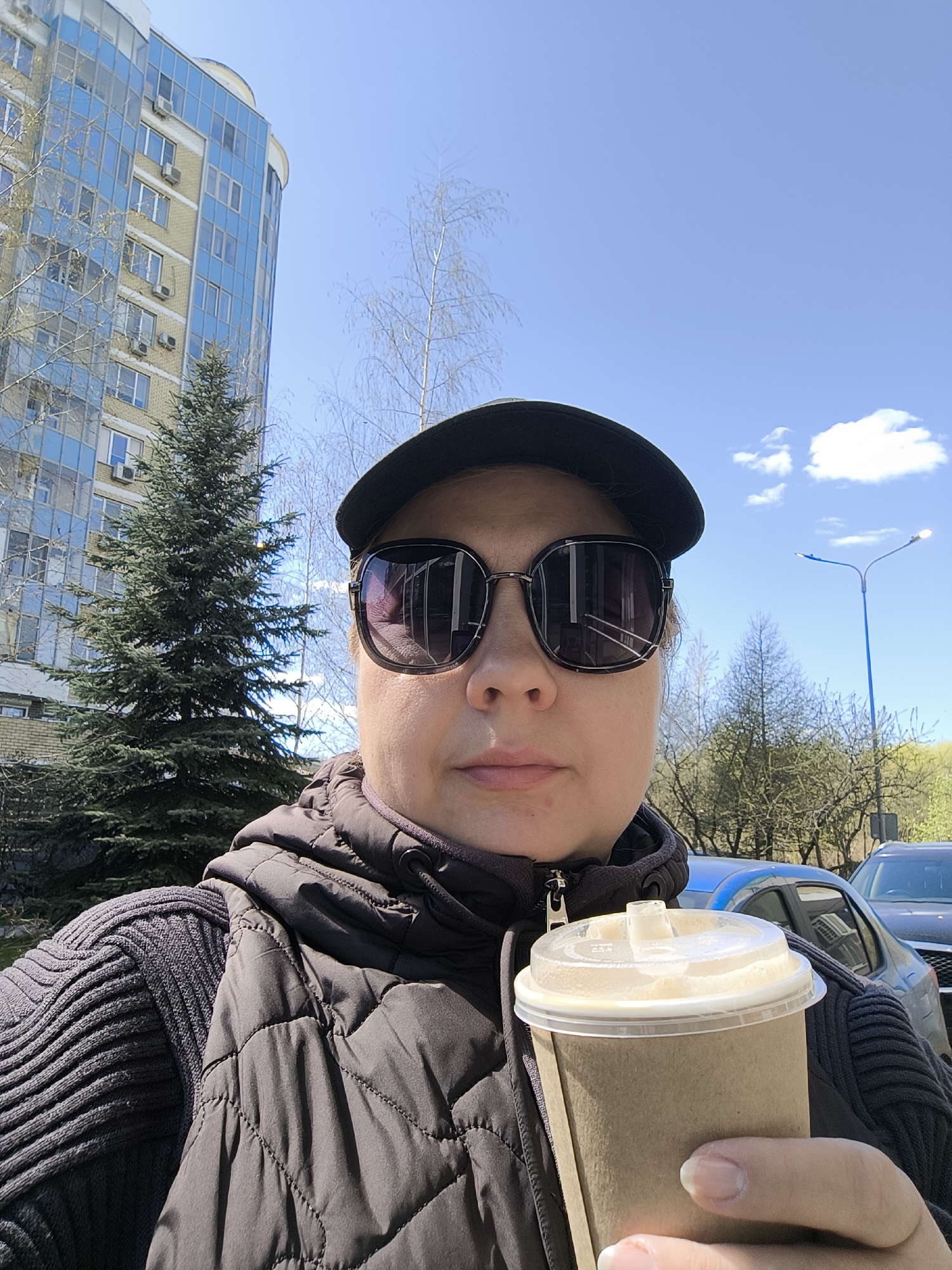 A middle-aged woman takes a selfie outdoors. She is wearing sunglasses, a dark cap, a quilted vest over a dark ribbed sweater, and holding a paper cup with a plastic lid, likely containing a drink. Behind her, there is a multi-story building with blue windows, a pine tree, other deciduous trees, a blue sky with some clouds, a street lamp, and several cars. The architecture suggests a location in a modern urban area, which appears to be Moscow, Russia.