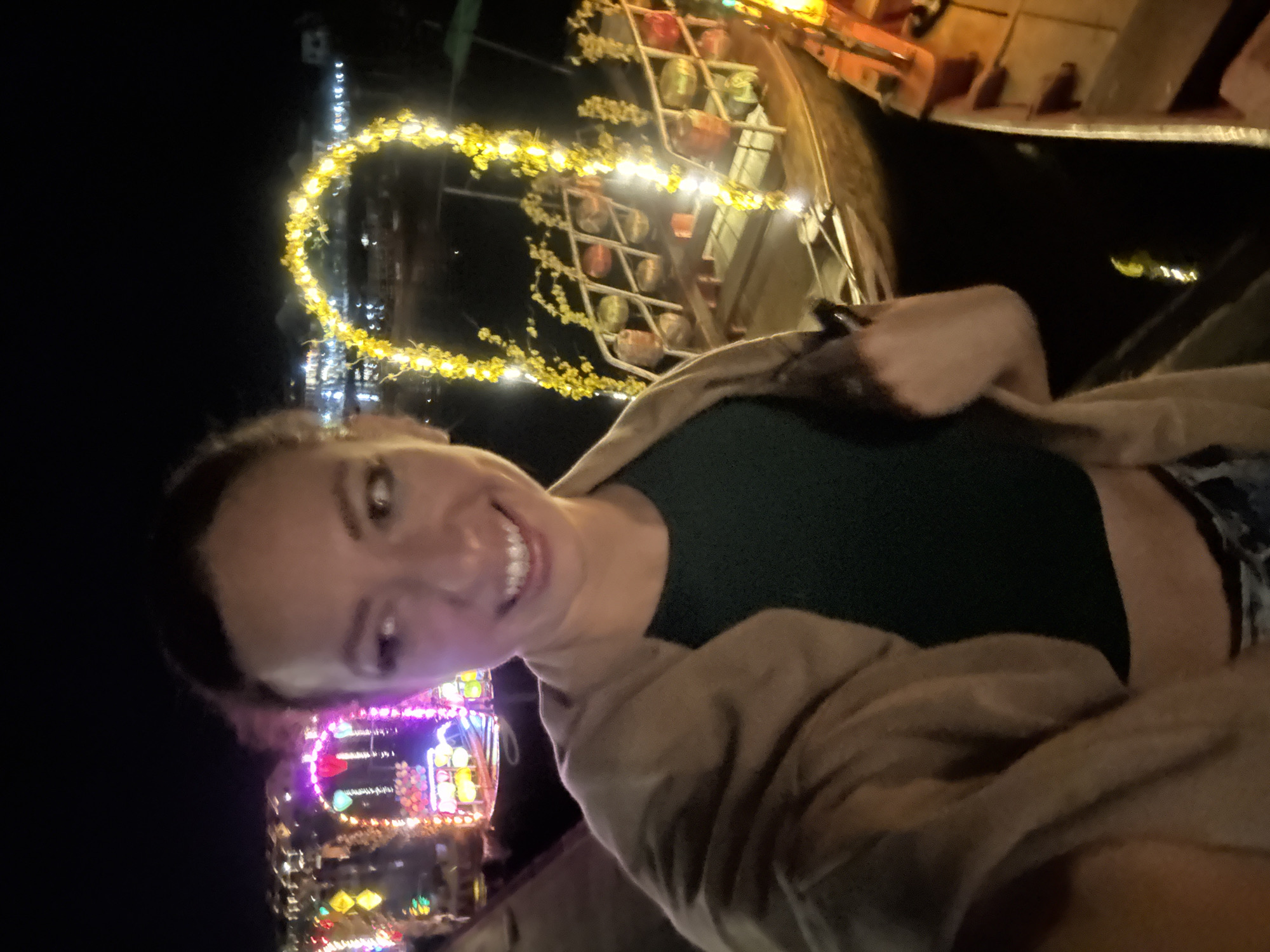 A young woman is taking a selfie in Hội An Ward, Vietnam, at night. The woman has fair skin, brown hair pulled back, and a wide smile revealing her teeth. She wears a dark green top, a light brown shawl or jacket, and what appears to be patterned shorts. In the background, there are light displays. Behind the woman and slightly to the left is a tower of bright, colorful lights. Above and to the right, a structure adorned with rows of lanterns and a curved line of yellow lights are visible. The overall setting suggests a festive or tourist-oriented environment. The image appears to be taken at a slightly tilted angle.
