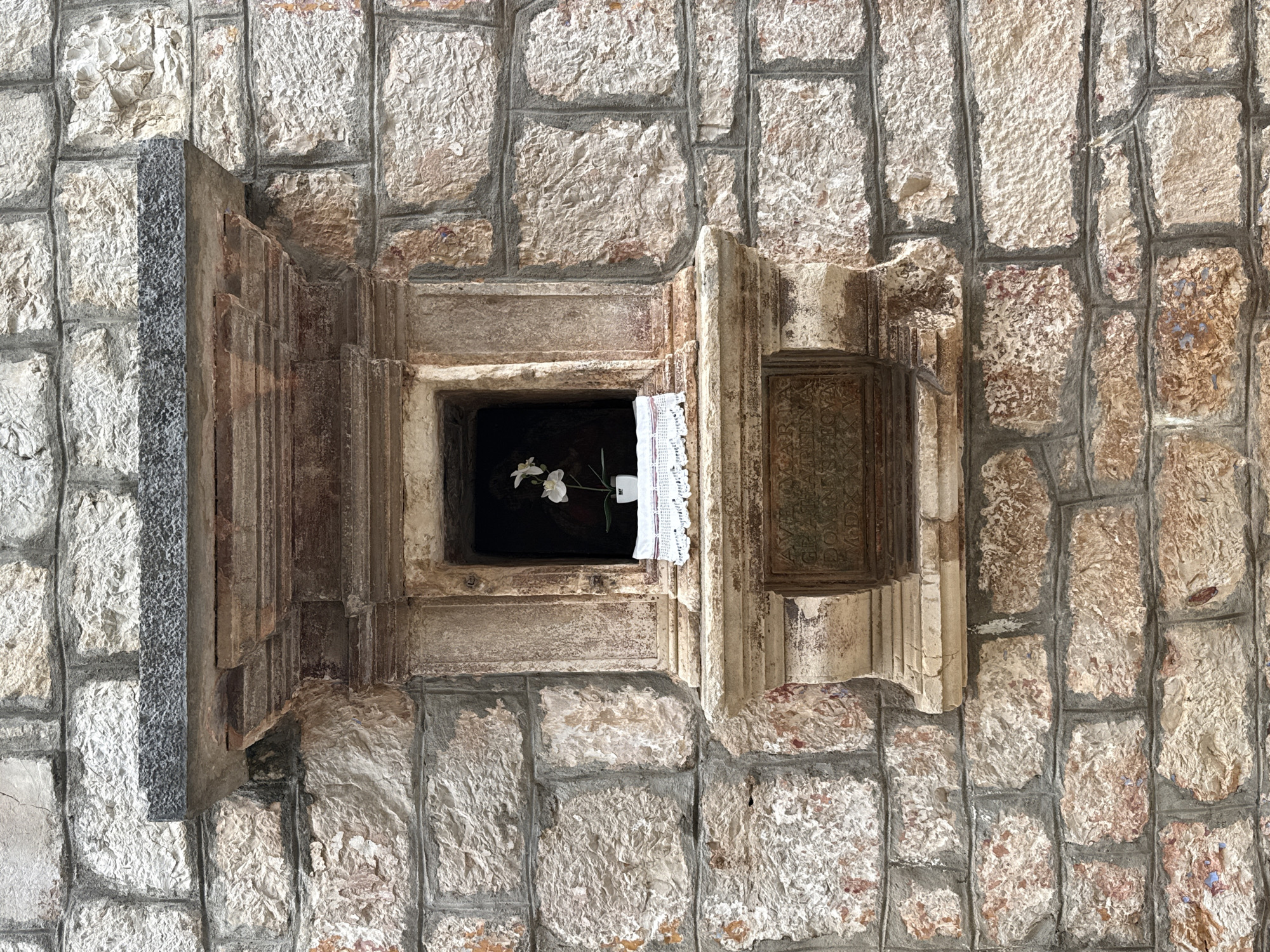 A small, ornate stone niche is set into a rough-hewn stone wall, characteristic of the architecture in the old town of Grad Stari Grad, Croatia. The niche features a small white orchid in a white pot placed on a white crocheted cloth. Above the niche, a dark, textured stone slab juts out, supported by carved stone brackets. Below the niche is a weathered bronze plaque with engraved text. The overall scene suggests a devotional space integrated into the historic urban fabric.