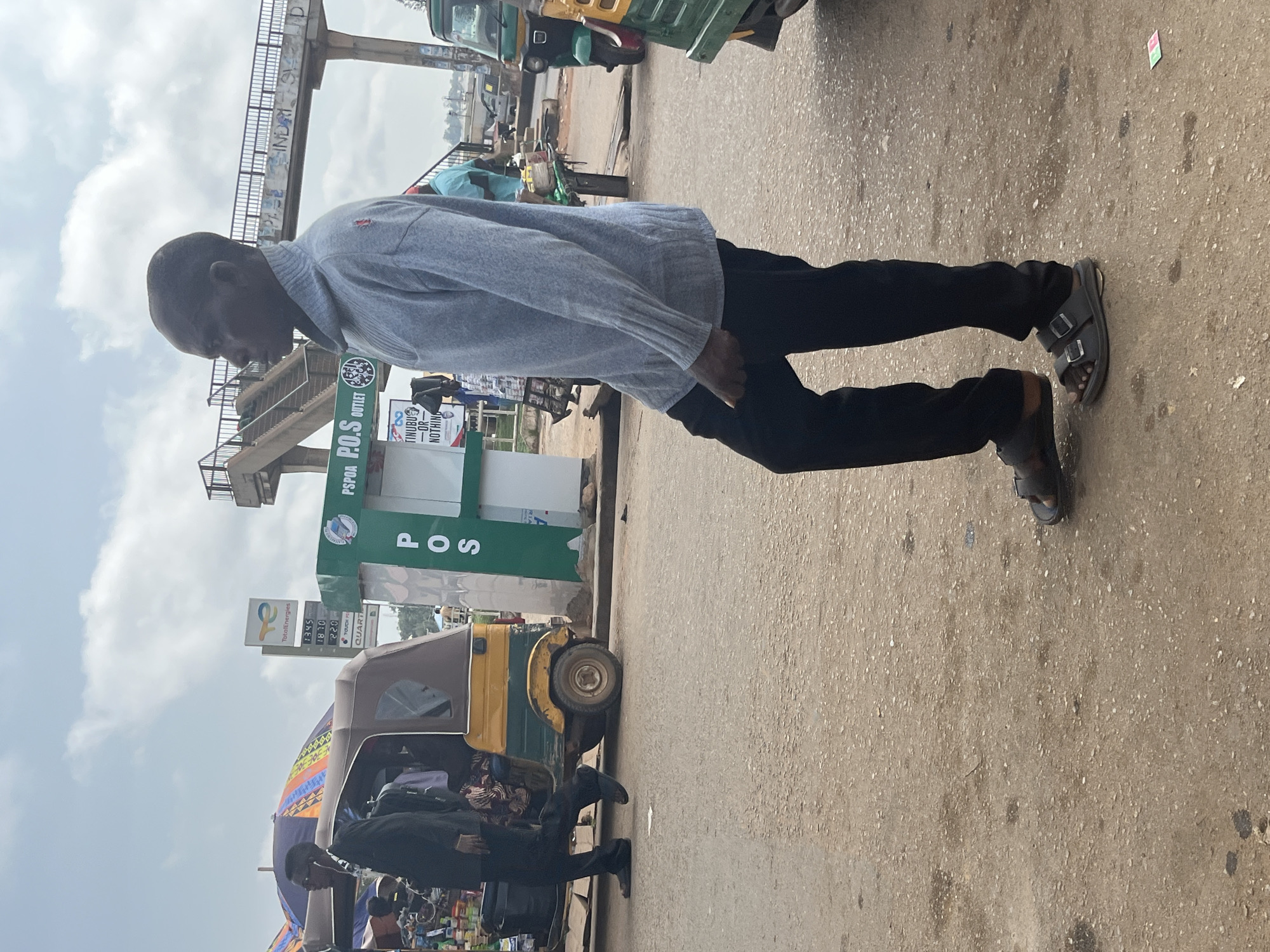 A man in a light blue sweater and black pants walks on a dusty road in Gura Top, Nigeria. He is wearing black sandals. To his left, a yellow and brown auto-rickshaw is parked. Behind the auto-rickshaw, a sign for "TotalEnergies" is visible, along with fuel prices. To the right of the auto-rickshaw, a green kiosk with "P.O.S OUTLET" signage is present. In the background, a pedestrian overpass spans the road. The sky is overcast.