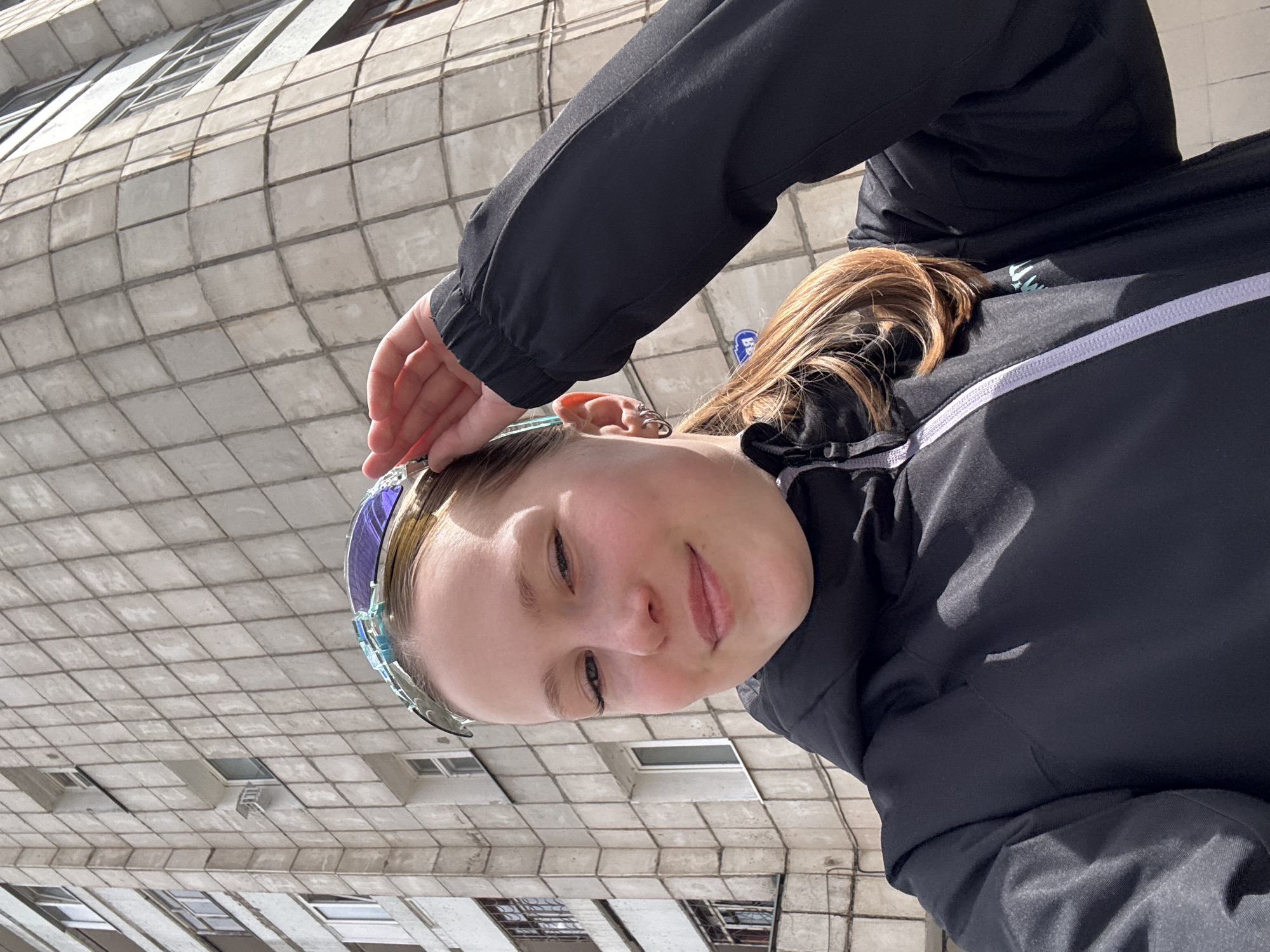 A selfie of a young woman in front of an apartment building, possibly in Chaikovsky, Russia. She is wearing a dark-colored jacket and glasses, and her hand is near her forehead. The building behind her is a high-rise apartment building with a tiled exterior and visible windows. The sky appears overcast.