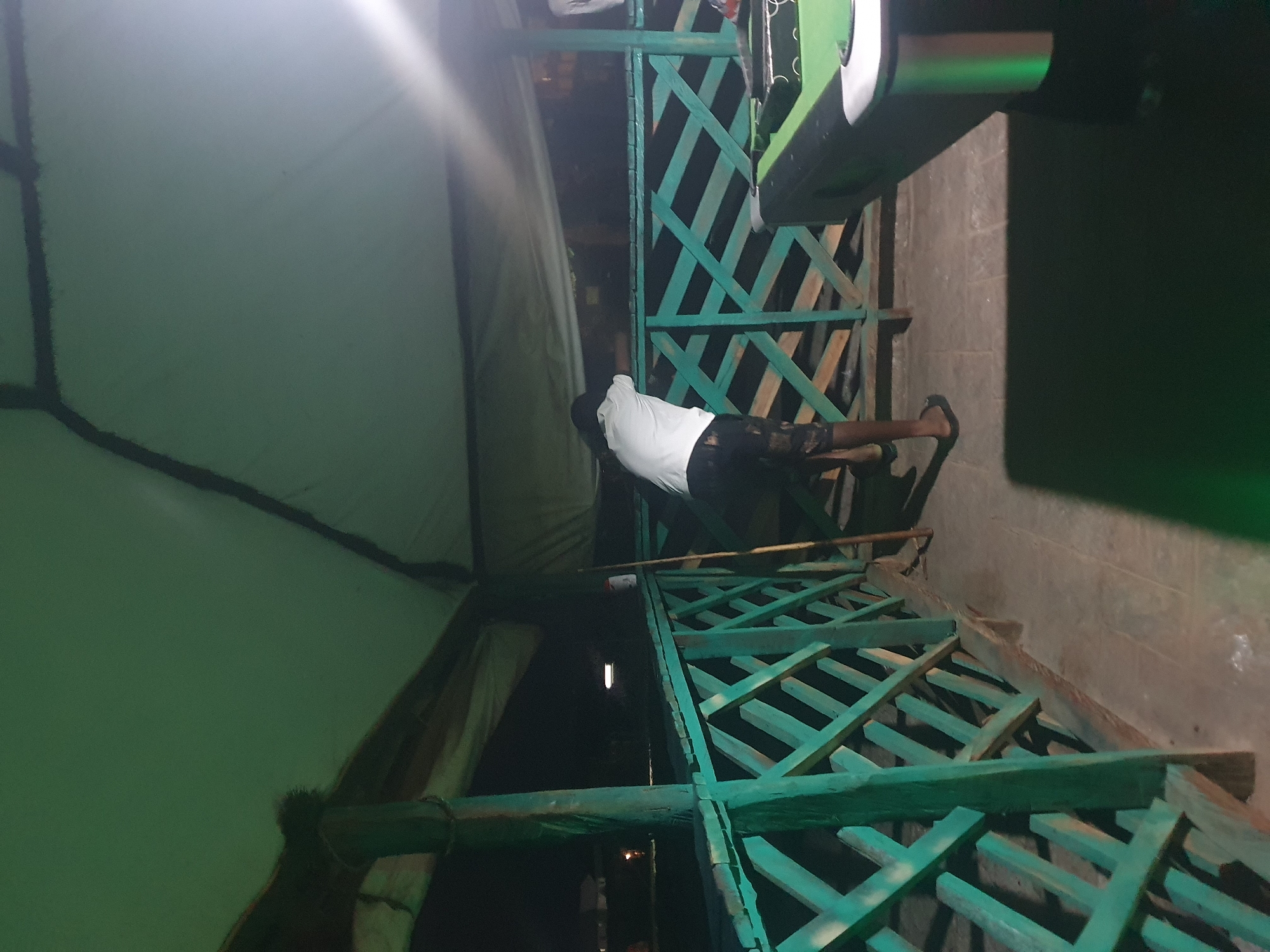 In Lokoja, Nigeria, a person wearing a white t-shirt and patterned shorts stands near a teal-colored wooden railing under a green canvas roof. The person is facing away from the camera, looking downwards towards a pool table visible on the right side of the frame. The pool table is green with black accents and illuminated by green lights. The environment appears to be an outdoor covered area, possibly a bar or recreational space, with wooden posts supporting the roof structure. The ground is paved with square tiles. In the background, the night sky is visible, with faint lights suggesting distant structures or streetlights. A long wooden pole leans against the railing to the left of the person.