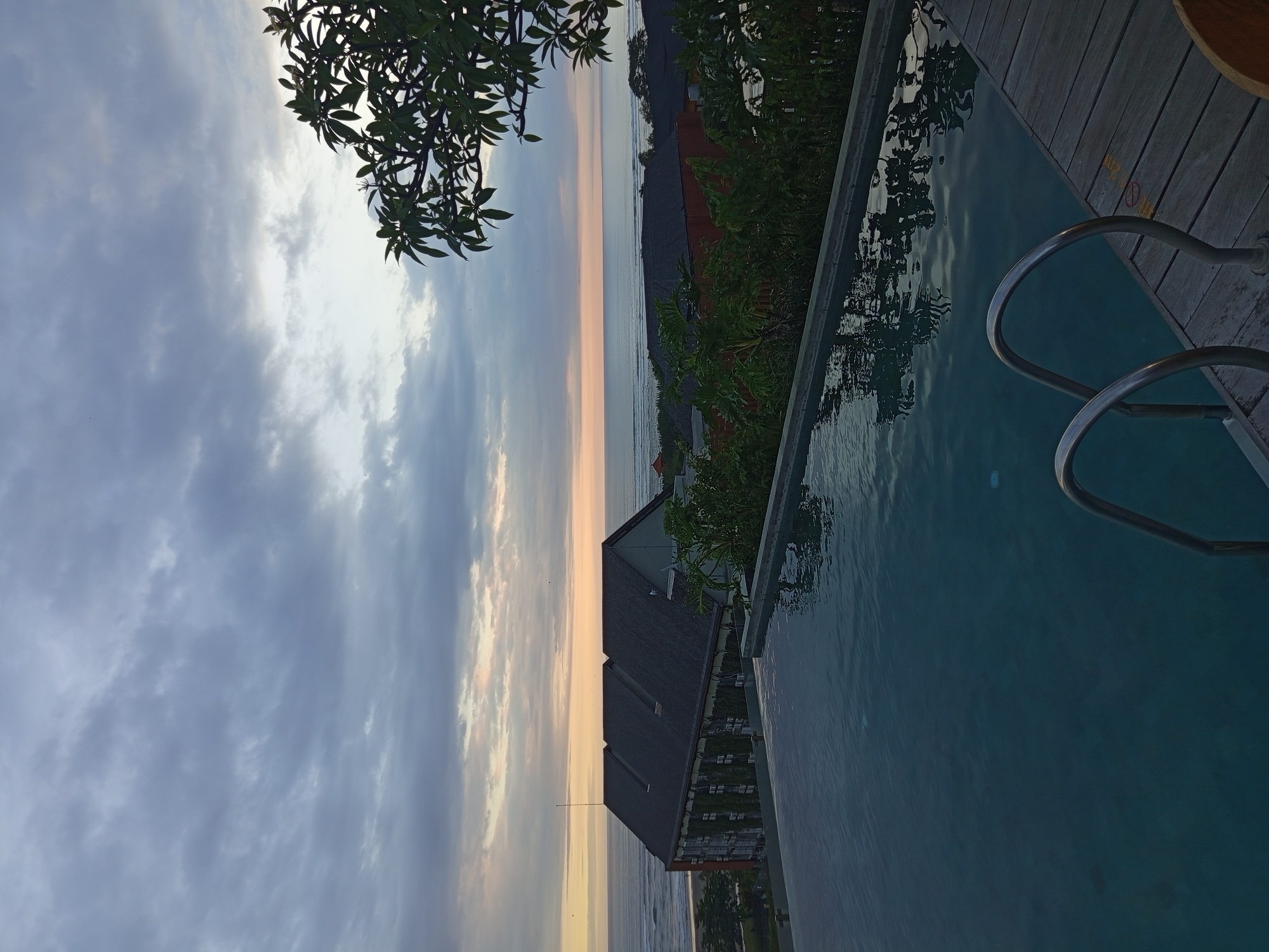 Captured from a high vantage point in North Kuta, Indonesia, the image presents a view encompassing a swimming pool, adjacent structures, and a distant seascape under a cloudy sky. The swimming pool, positioned on the right side, has a ladder, and its waters reflect the surroundings. A wooden deck borders the pool. Moving toward the center, the roofs of buildings, likely accommodations, are visible. Vegetation is interspersed among the structures. In the distance, the ocean meets the horizon under a sky filled with clouds. The light suggests either sunrise or sunset. The frame is also partially occupied by tree branches in the upper left corner.