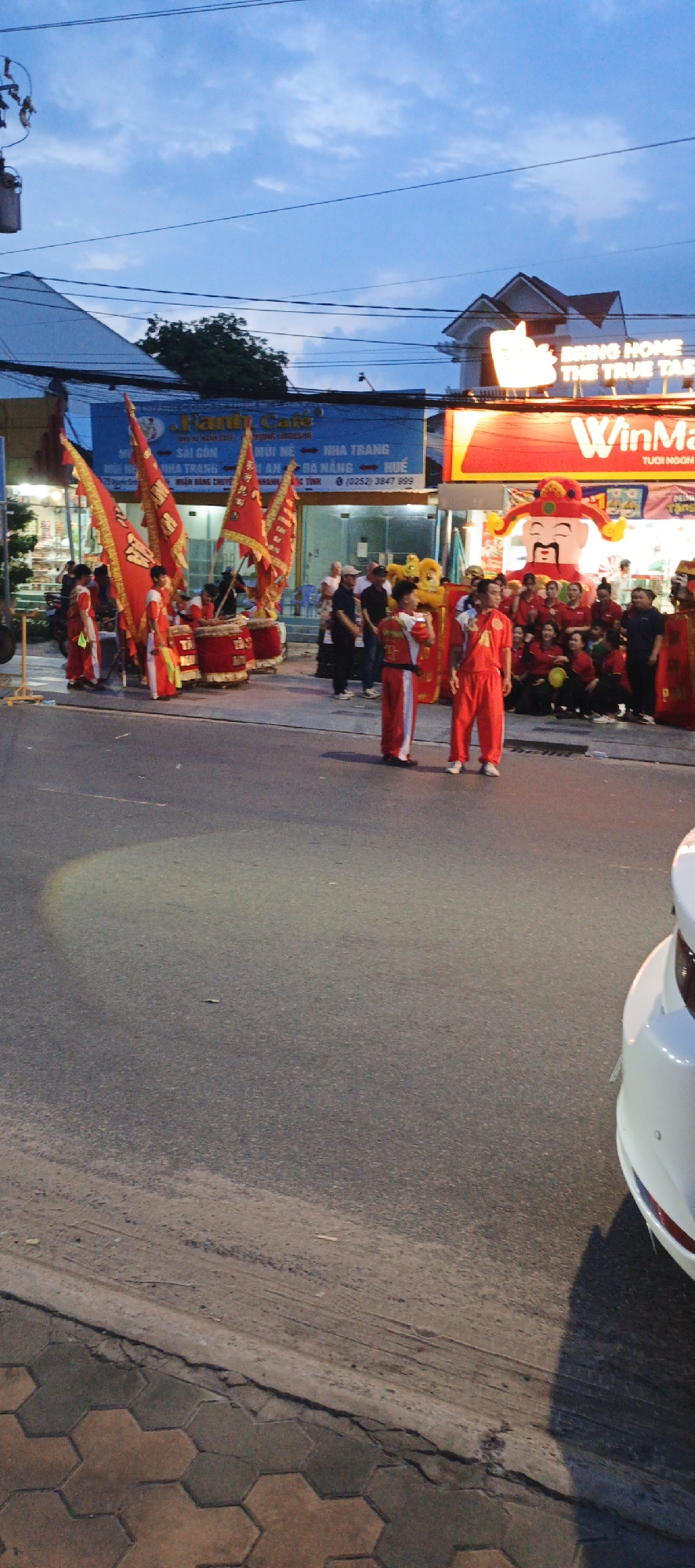This image captures an outdoor street scene at dusk in Unknown, Unknown, likely in Southeast Asia (specifically Vietnam, given the Vietnamese text on signs). The foreground features an asphalt road with a segment of a hexagonal-tiled sidewalk on the left.
The central focus is a cultural performance or celebratory event. On the left, several individuals in red and white traditional costumes are positioned around large red drums and multiple prominent red flags adorned with gold embroidery. Two young males in identical red and white attire stand centrally on the road, facing right, observing the event.
To the right, a WinMart supermarket is prominently displayed with its brightly lit red signage. In front of WinMart, a large inflatable red figure resembling a smiling deity with a traditional hat stands as a focal point. A yellow lion dance mascot is also visible near the entrance. A group of approximately 10-15 people, many wearing red shirts, are gathered near the WinMart entrance, some crouching, others standing, observing the scene. Additional casually dressed individuals are scattered throughout the background.
Overhead, numerous electrical wires traverse the sky, which displays a twilight hue with visible clouds. A blue-painted building with a sign (partially readable as "Family Cafe" and listing Vietnamese cities like "SÀI GÒN," "MŨI NÉ," "NHA TRANG") stands to the left of WinMart. A residential-style building with a peaked roof is visible in the background above the WinMart sign. A white car is partially visible on the far right of the frame.