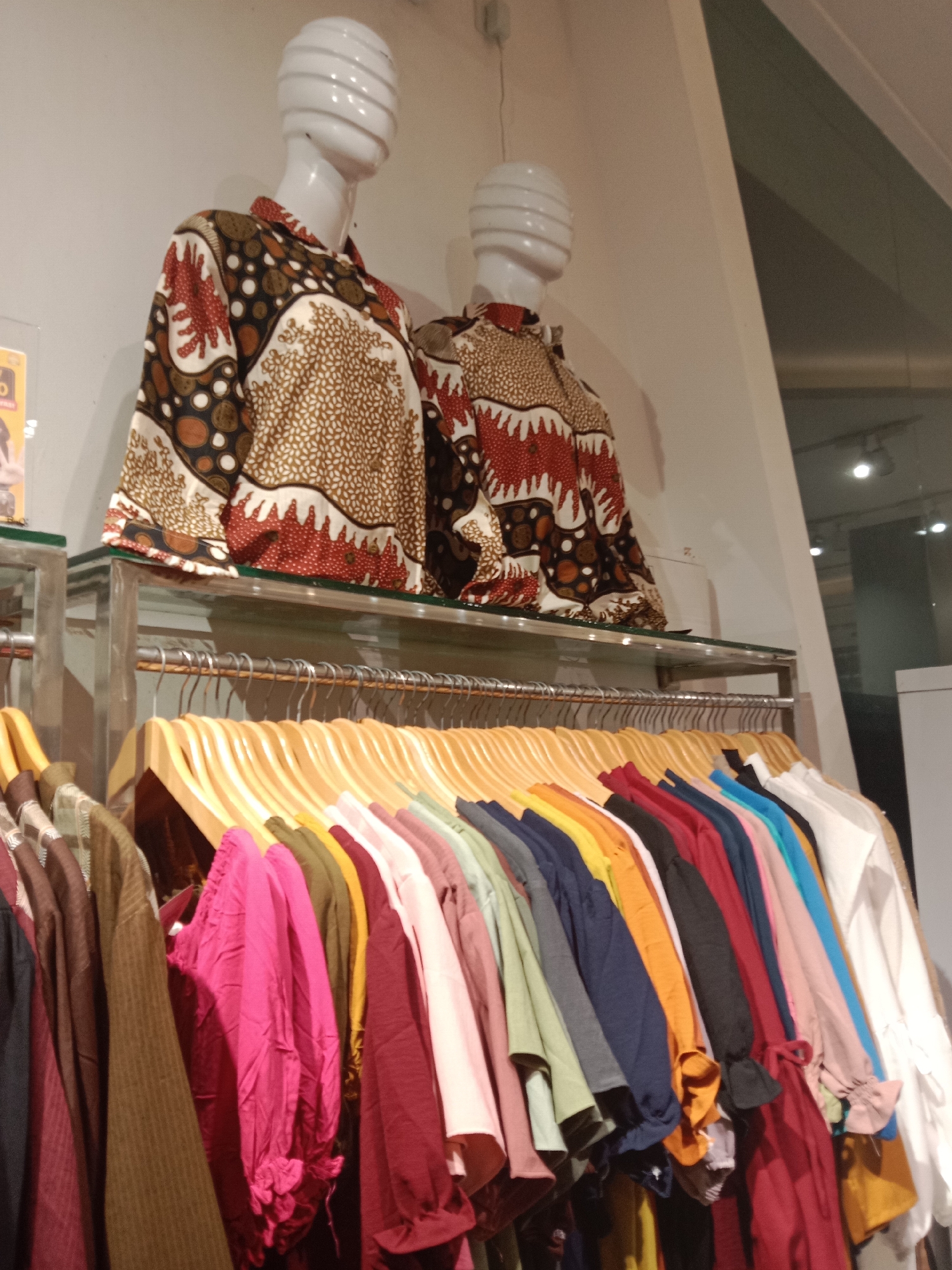This is a medium shot, slightly angled, indoor photograph of a clothing display inside a store. Two mannequins, positioned side-by-side and facing left, are wearing patterned batik shirts. The mannequin on the left wears a shirt with a brown, cream, and red pattern featuring circular and wave-like motifs. The mannequin on the right wears a shirt with a red and white wavy pattern. Both mannequins have white, ribbed heads.
Below the mannequins, a horizontal rack holds numerous shirts on wooden hangers. The shirts display a vibrant spectrum of colors, including pink, maroon, olive green, mustard yellow, orange, dark blue, black, light blue, and white. The lower part of the shirts are visible, showing various sleeve styles and details such as ruffles.
The background consists of a plain, off-white wall and a dark ceiling. To the right of the mannequins, a large pane of glass reflects some of the interior lighting and a hint of an outdoor scene. The store appears to be a retail environment, likely a clothing boutique or department store.
The image was taken in Semarang, Indonesia.