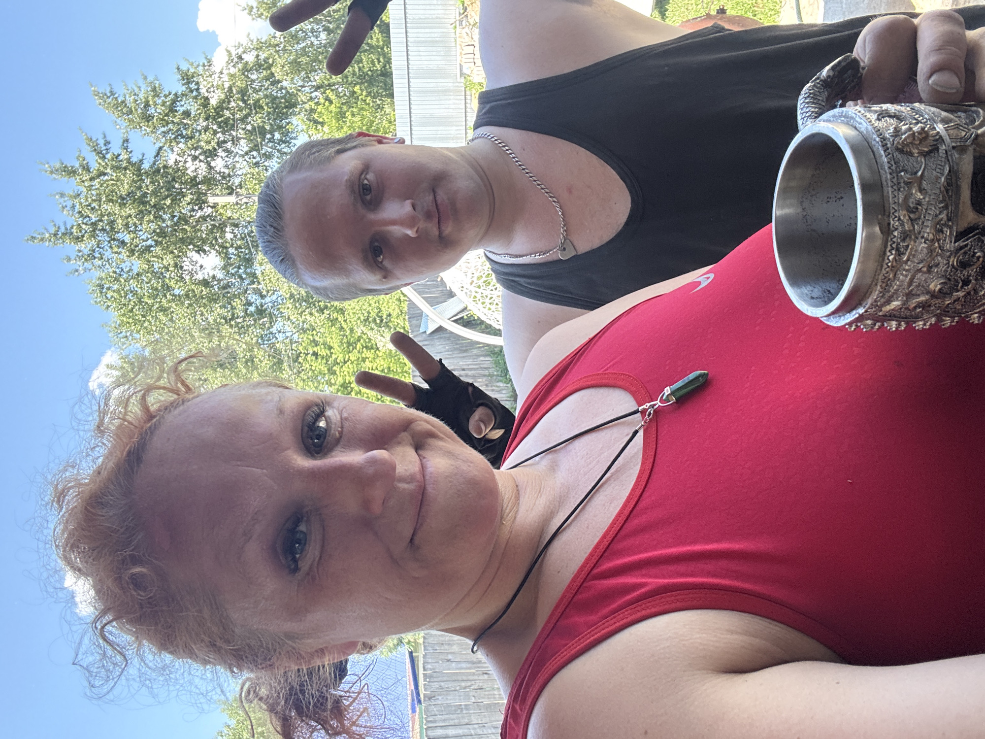 A selfie shows a Caucasian woman and a Caucasian man outdoors on a sunny day. The woman, in the foreground and on the left, has curly red hair and is wearing a red tank top and a necklace with a green pendant. Her right hand is raised, with two fingers extended in a peace sign, and she is wearing fingerless gloves. She is smiling. The man is to her right, wearing a black tank top and a silver chain necklace with a heart pendant. His left hand is also raised, with two fingers extended in a peace sign, and he is wearing fingerless gloves. He is holding a decorative metal mug with a snake-like handle in his right hand. In the background, there is a wooden fence, green trees, and a blue sky with scattered clouds. The presence of a wooden fence and the style of housing suggest a dacha or a private residential area common in Russian cities like Chaikovsky.