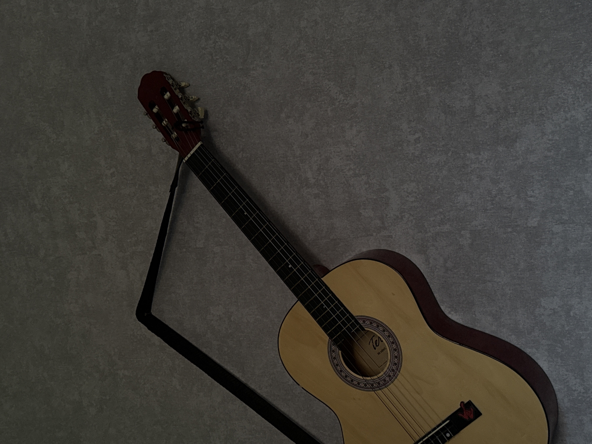 The image shows a classical guitar with a dark strap draped over it, leaning against a textured, gray wall. The guitar is light wood with a red headstock and dark fretboard. The lighting is dim, casting shadows and highlighting the grain of the wall and the guitar's body. There are no people or animals present. The image appears to be taken indoors. No information about the location of Abinsk, Russia, can be inferred from the visual content of the image.
