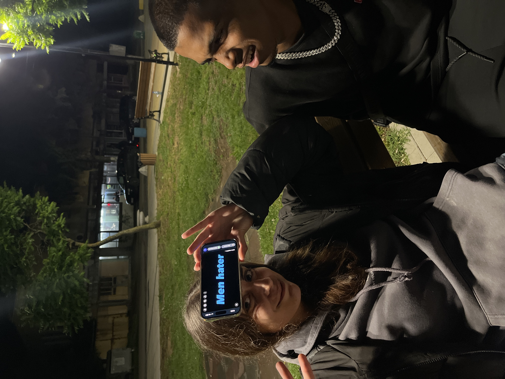 The image captures two individuals outdoors at night in what appears to be a park or urban area in Lozarevo, Bulgaria. A young woman with long dark hair is in the foreground, seen from the chest up, looking at the camera with a slight smile or smirk. She wears a dark jacket or hoodie and holds a smartphone in her left hand, displaying its screen. With her right hand, she forms a peace sign. The phone screen prominently displays the text "Men hater" in bright blue.
To her upper right, a young man is partially visible, from the shoulders up, with short dark hair and wearing a dark top adorned with a silver chain. He looks down towards the woman or the phone, appearing engaged with a subtle smile.
The background shows a dark, nighttime setting with artificial lighting. A street light illuminates a leafy tree on the left, and a paved path runs through a grassy area. A wooden bench is visible, along with glimpses of a building with lit windows and a parked car further in the distance. The scene suggests a casual, playful interaction between the two subjects during the evening.
