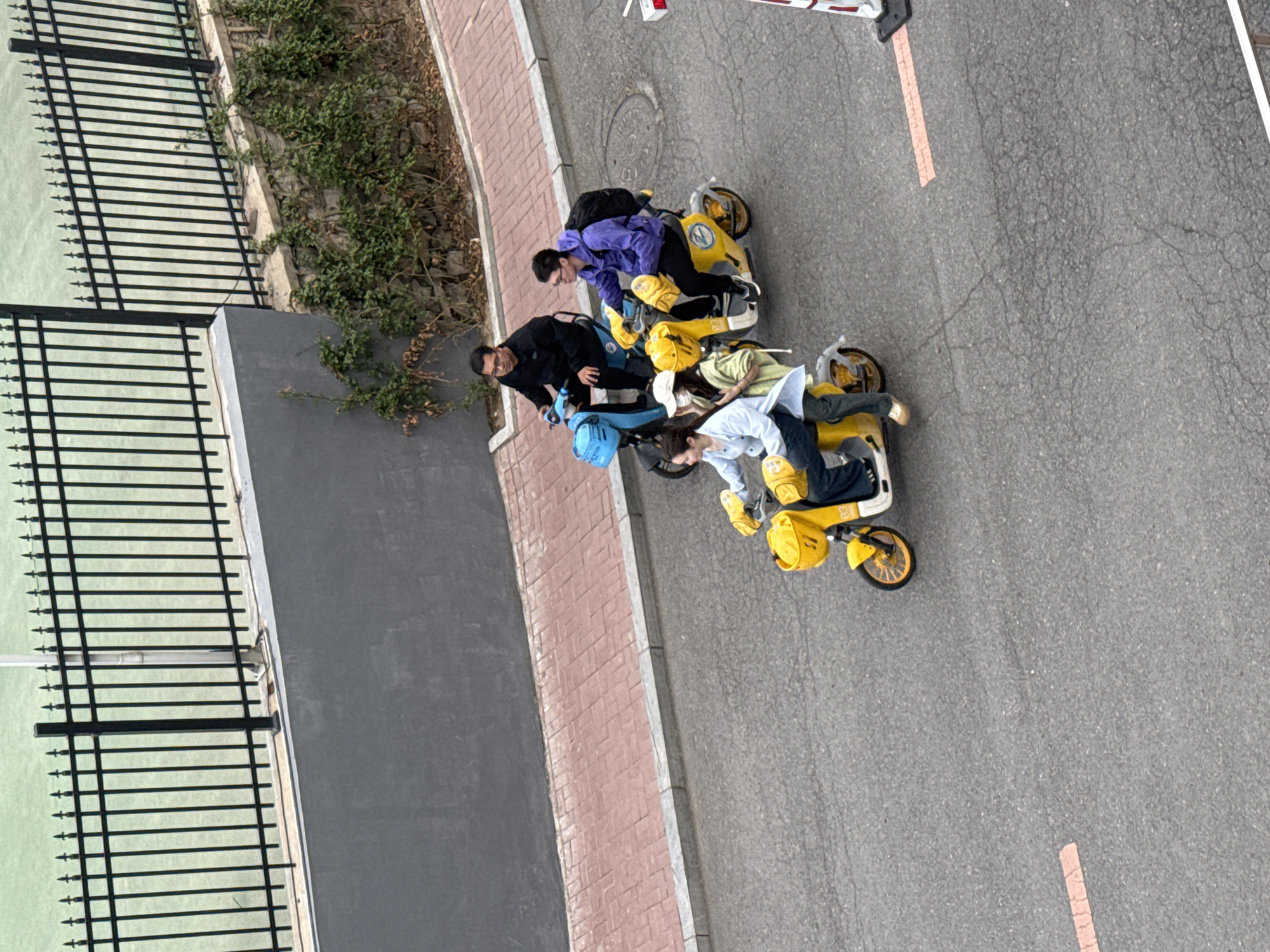 This high-angle image, set in Zhongtun, China, captures four individuals interacting with three shared electric scooters on an asphalt road. The scene takes place during a clear day, indicated by natural lighting.
In the foreground, a group of four young adults is clustered around the scooters. To the left, a person in a black jacket leans over a light blue electric scooter, which has a matching blue basket. Adjacent to them, another individual wearing a purple jacket and black backpack is positioned over a yellow electric scooter, featuring a "U" logo on its front basket and yellow footrests. Further right, a person in a light green top and white cap is lying horizontally across the seat of a yellow scooter. Next to them, another individual in a light blue shirt and dark pants is similarly stretched across a second yellow scooter. This yellow scooter has the number "85" visible on its side. Their unusual poses suggest playful interaction or posing for a photograph.
The scooters are parked on a grey asphalt road that shows minor cracking. A reddish-brown brick sidewalk runs parallel to the road, bordered by a low concrete wall and a small section of wild green foliage. To the far left, a tall black metal fence encloses an area with a light green surface, possibly a tennis court or other sporting ground. A white object with red markings, likely part of a barrier or sign, is partially visible in the upper right corner of the frame. No explicit text other than the scooter markings is clearly discernible.