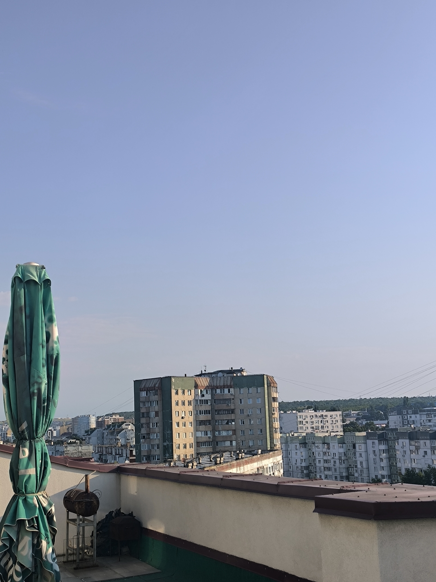 A rooftop view overlooking a residential area in Chișinău, Moldova, is captured under a clear blue sky. In the foreground, a green, folded umbrella with a patterned design stands prominently on the left. To its right, a rusty barbecue grill sits on a metal stand, with a pile of dark material, possibly charcoal or wood, next to it. A wall separates the rooftop area from the city view, with a red trim along its top.
Beyond the rooftop, a cluster of multi-story apartment buildings dominates the middle ground. One prominent building in the center is tall and has a greenish-brown facade with numerous windows. To its right, a row of lighter-colored apartment buildings extends into the distance. Further back, more buildings and a dense line of trees are visible, suggesting a larger urban landscape. Power lines are also strung across the scene in the background. The lighting indicates it is likely daytime, with the sun casting a gentle glow on the buildings. There are no people visible in the image.