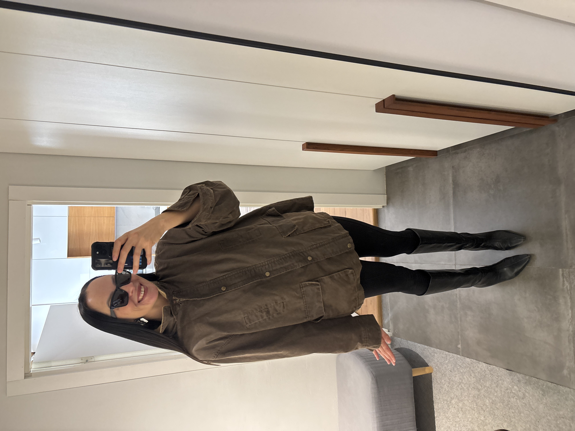 This image, taken in Chaikovsky, Russia, depicts a female subject in an indoor setting, likely a dressing room or hallway, captured via a selfie reflected in a mirror. The subject is a woman with long, dark hair, wearing dark rectangular sunglasses and light-colored, geometric earrings. She is smiling, revealing her teeth. Her attire includes a loose-fitting, medium-brown or olive-drab jacket with a zippered front, a collar, and multiple flap pockets secured with buttons. She wears tight black leggings or pants and black, pointed-toe, knee-high boots. Her right hand holds a dark smartphone, positioned to take the photograph, while her left hand is extended outward and slightly upward to her left, palm open.
The immediate environment includes a dark gray tiled floor. To the subject's left, a gray upholstered bench or ottoman with light wooden legs is visible. The background, reflected in the mirror, shows light-colored walls. An opening on the left side reveals a brighter room beyond with light wooden surfaces and a ceiling light fixture. To the subject's right, a light-colored wardrobe or cabinet structure with two vertical, rectangular wooden handles is present. The mirror's dark frame is partially visible on the far right edge of the image.