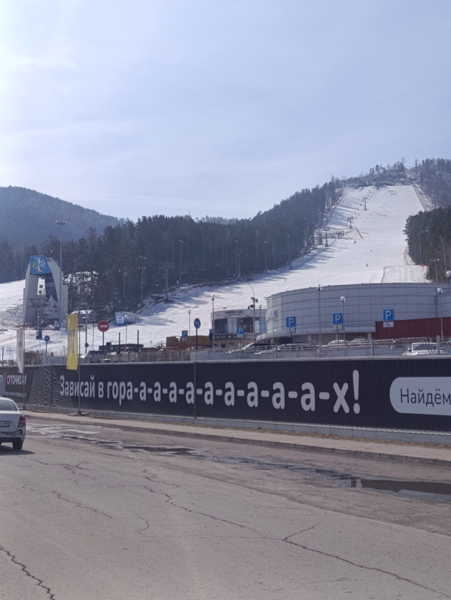 The image shows a street scene outside the Bobrovy Log ski resort in Krasnoyarsk, Russia. The main focus is on a long black advertising fence with white text in Russian: "Зависай в гора-а-а-а-а-а-а-а-а-а-х!" and "Найдём". The fence runs parallel to a cracked asphalt road with puddles. Behind the fence is the ski resort, featuring snow-covered ski slopes, ski lifts, a large modern building with "P" signs indicating parking, and trees. The sky is overcast and pale blue. A car is visible on the left side of the image. The lighting suggests it is daytime.