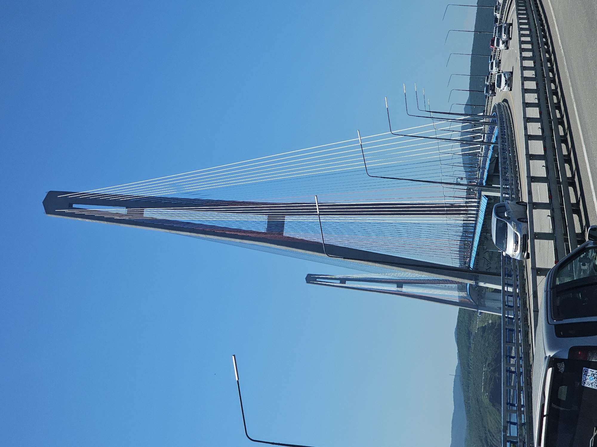 The image shows a view of the Russky Bridge in Vladivostok, Russia, on a clear, sunny day. The bridge, a prominent landmark, is a cable-stayed bridge known for its impressive height and modern design. Its two tall pylons, which taper towards the top and are connected by a network of white cables, dominate the skyline. The road deck of the bridge is visible, with several cars driving on it. The scene is framed by a bright blue sky and rolling, tree-covered hills in the background, suggesting a coastal or island environment. Streetlights with sleek, modern designs are spaced along the roadway, illuminating the bridge's path. The overall impression is one of a well-maintained, significant piece of infrastructure set against a natural, scenic backdrop.