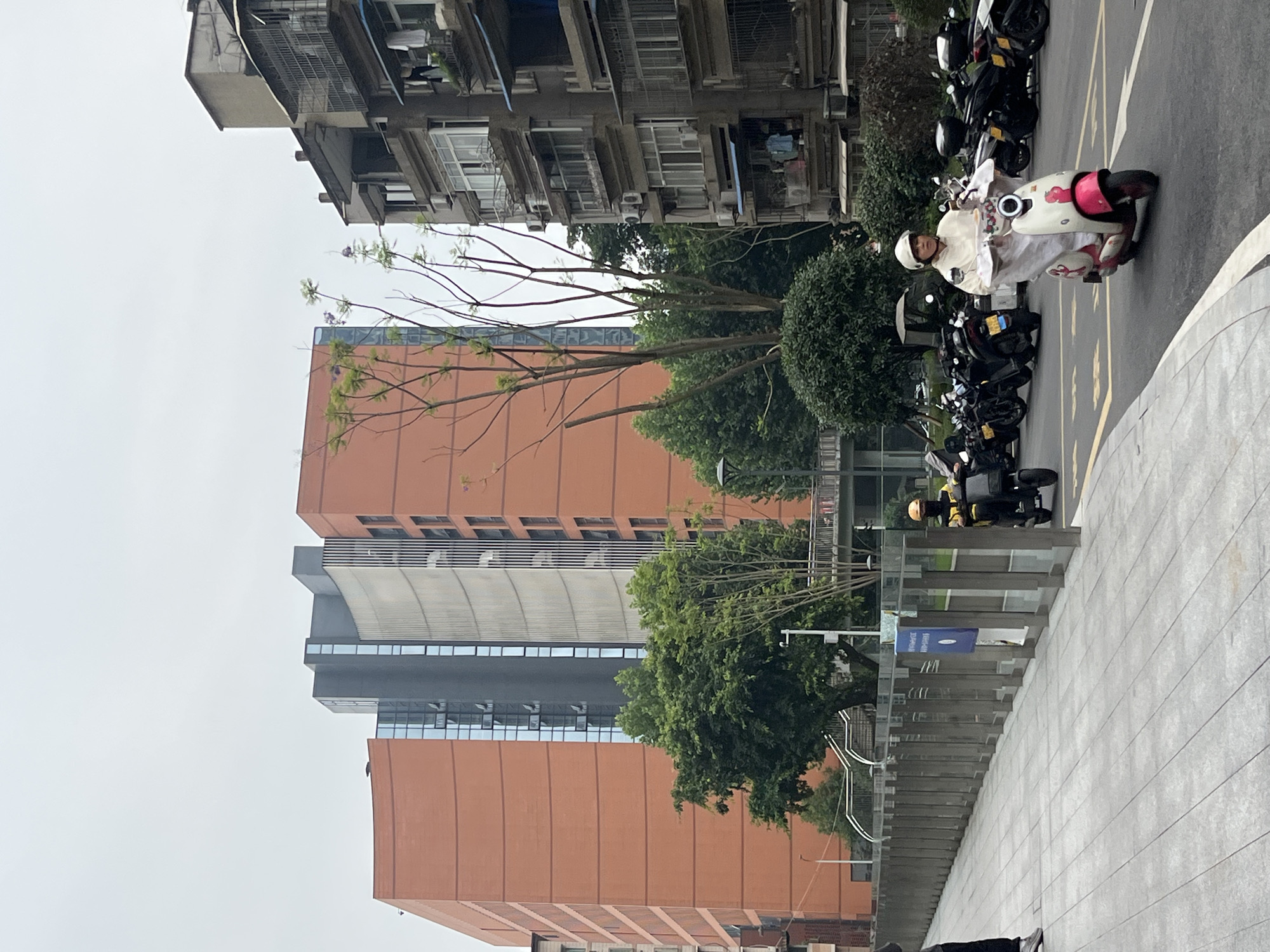 A medium shot shows a street scene in Yuzhong District, China. In the foreground, a person rides a pink and white scooter. The rider is wearing a white hooded jacket and a white helmet. The scooter has a cartoonish design on its front. To the left, another scooter is partially visible with a rider in a yellow jacket and helmet. Further back, a row of parked scooters lines the street. The background features a modern, orange-brick building with dark, glass-paneled sections. To the right of this building, an older, multi-story residential building with balconies and air conditioning units is visible. Trees and foliage are interspersed throughout the scene. The sky is overcast.