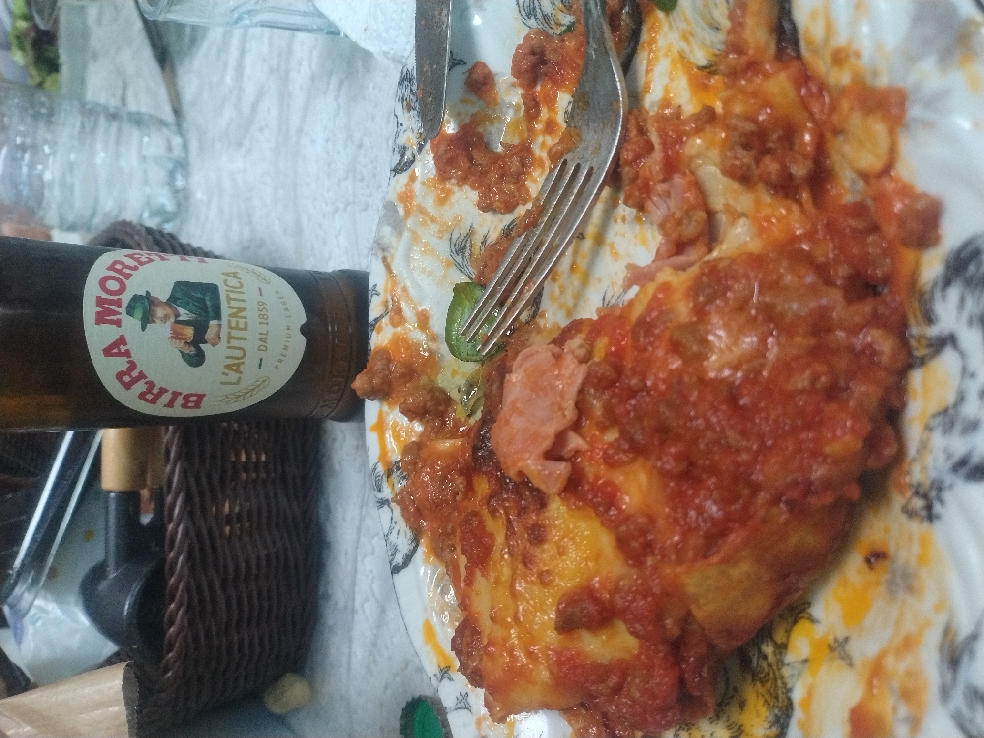 The image shows a plate of lasagna with a fork and knife lying on top. Next to the plate is a bottle of Birra Moretti. There is also a wicker basket and a water bottle visible in the image. The setting appears to be indoors, possibly a home or restaurant in Luxembourg, Luxembourg, given the presence of Birra Moretti, which is often found in Italian restaurants and homes.