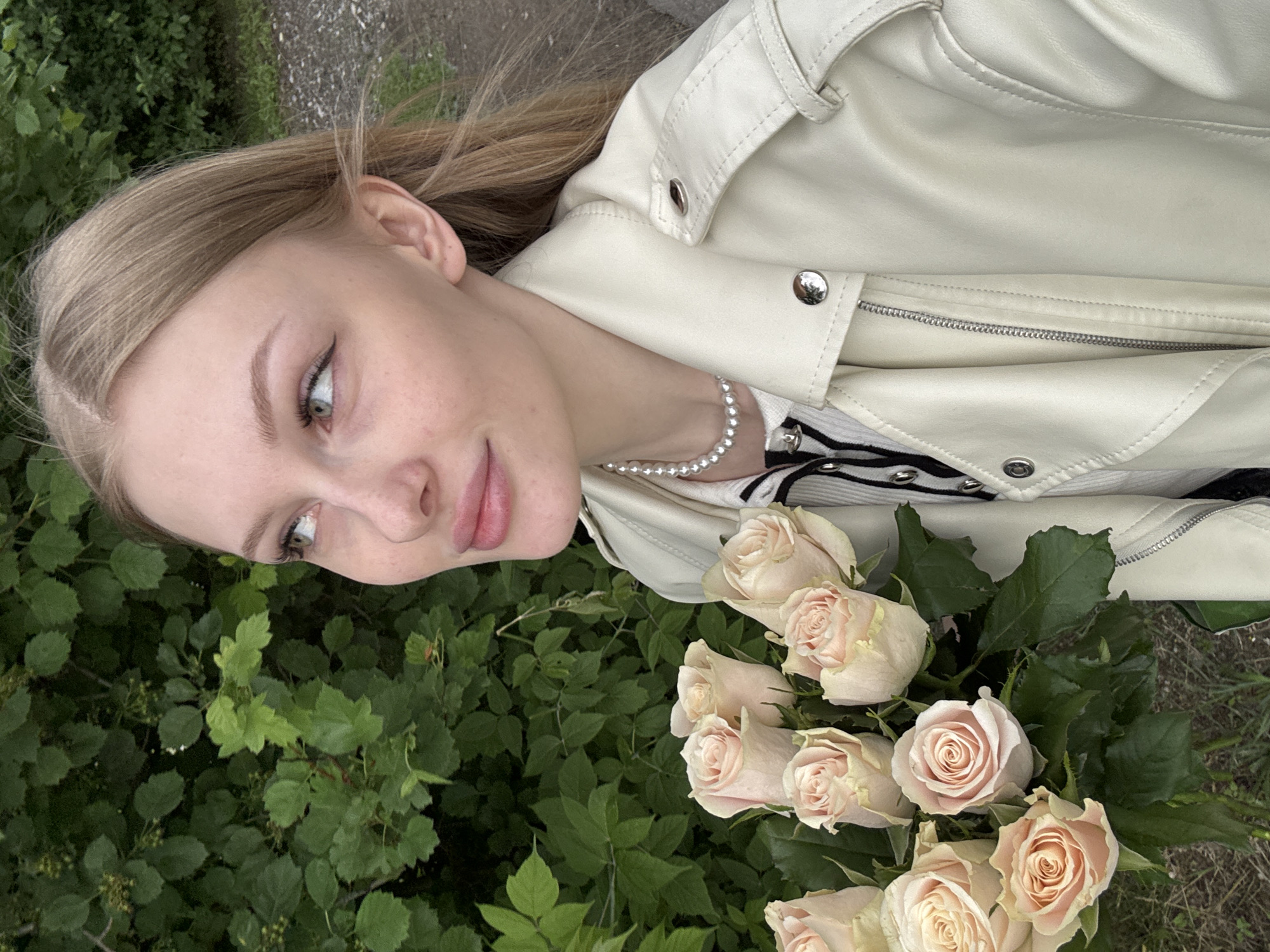 A young, fair-skinned woman with long, light-blonde hair is depicted outdoors, holding a large bouquet of light pink roses. She wears a cream-colored faux leather moto-style jacket, featuring silver snaps on the lapels and shoulder straps, and a prominent front zipper. Underneath, she has on a white ribbed top with black horizontal stripes and black buttons. A single strand pearl choker necklace is visible on her neck. Her light-colored eyes, lined with black eyeliner, are directed upwards and to the viewer's left, with a subtle, closed-lip smile. The background consists of dense, vibrant green foliage, suggesting a natural outdoor environment such as a park or garden. A portion of a gravelly path is visible to the right and at the bottom of the frame. This scene is located in the city of Perm, Russia.