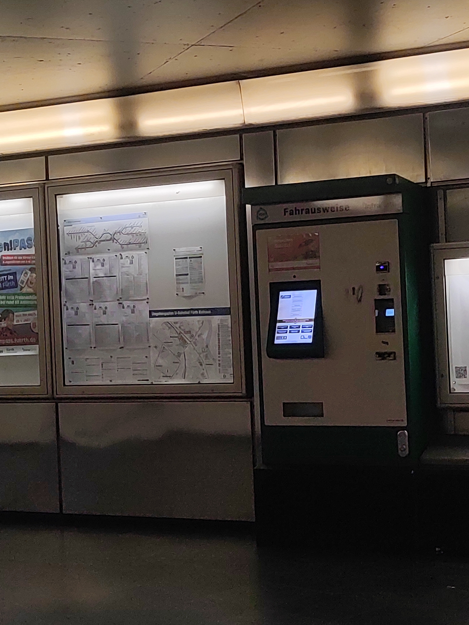 The image shows a public transportation ticket vending machine at the U-Bahnhof Fürth-Stadthalle in Fürth, Germany. The machine, labeled "Fahrausweise," is green and white and features a touchscreen display. To the left of the machine are display boards showing public transport routes and information, one of which has the text "eniPASS." To the right of the vending machine is another illuminated display case. The environment appears to be an underground station with a tiled wall and overhead lighting. The scene is devoid of people.