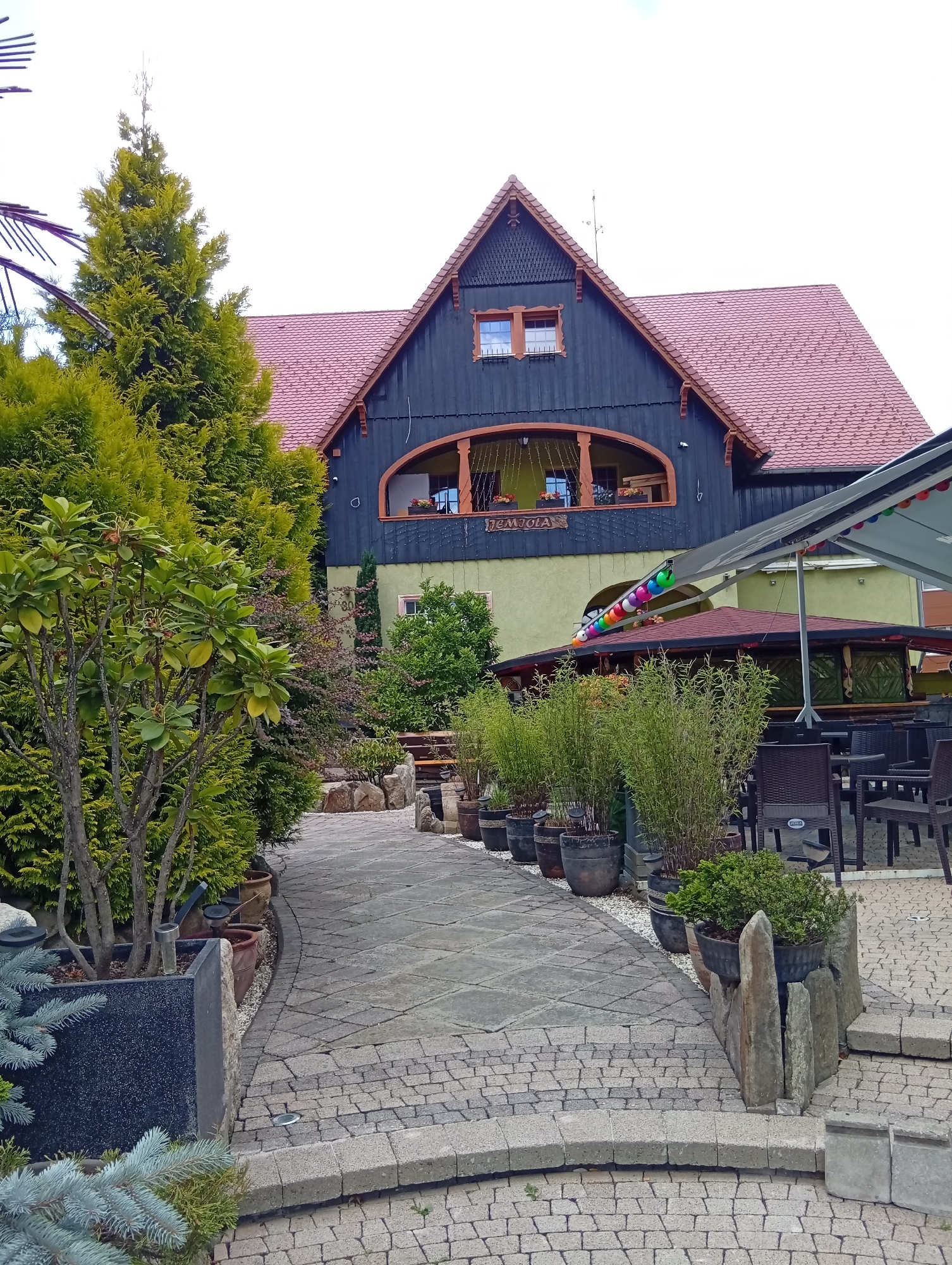 The image depicts an exterior view of a building with a distinctive dark wooden upper story and a light green lower story, topped with a reddish-brown tiled roof. A large, triangular gable faces the viewer, featuring decorative wooden trim. The building has arched windows and a prominent balcony area. To the left, a tall, vibrant green coniferous tree stands next to a smaller, leafier tree with bare branches, planted in a large dark rectangular planter. A stone pathway paved with irregularly shaped stones leads towards the building, curving to the right. Along the pathway, various potted plants and stones are arranged. To the right, an outdoor seating area is visible, with dark chairs and tables beneath a retracted awning decorated with colorful balloons. A smaller, separate structure with a dark roof and open sides is situated in front of the main building, also featuring seating. The overall scene suggests a commercial establishment, possibly a restaurant or guesthouse, situated in a garden-like setting. The sign above the balcony on the main building reads "TEMJOLA". The presence of outdoor seating and decorative elements indicates a welcoming ambiance for patrons.