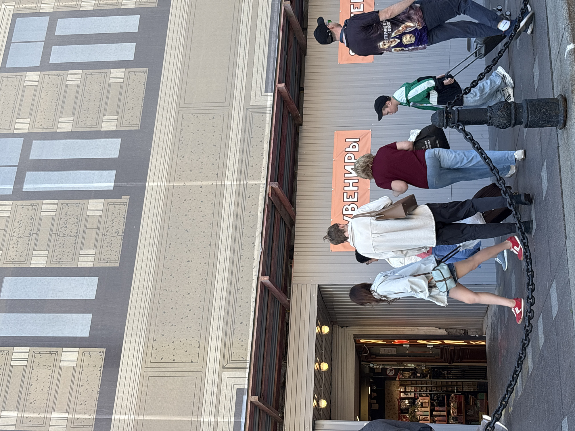 The image depicts a street scene in Saint Petersburg, Russia, in front of a building with a large mural of windows. Several people are walking on the sidewalk. In the foreground, a chain barrier runs across the sidewalk.
From left to right, a person wearing a dark jacket is partially visible. Next, a young woman with long dark hair, wearing a light blue jacket and denim shorts, walks with red sneakers. She is carrying a light blue purse. Behind her, a woman with blonde hair, wearing a maroon top and blue jeans, walks with a shopping bag. A person wearing a light-colored jacket is walking behind the blonde woman. Further right, a young boy in a green and white jacket is walking with a black rolling suitcase. To the far right, a man in a black cap and black t-shirt with an image of Stephen Curry is walking with a black rolling suitcase.
Two large orange banners are visible on the building wall. The left banner has Cyrillic text that appears to read "СУВЕНИРЫ" (Souvenirs). The right banner is partially obscured.
The building facade above the banners features a large, stylized mural of windows and architectural elements. The area below the mural appears to be an entrance to a shop, with a metal awning and lights visible. The overall impression is that of a commercial area or tourist spot.