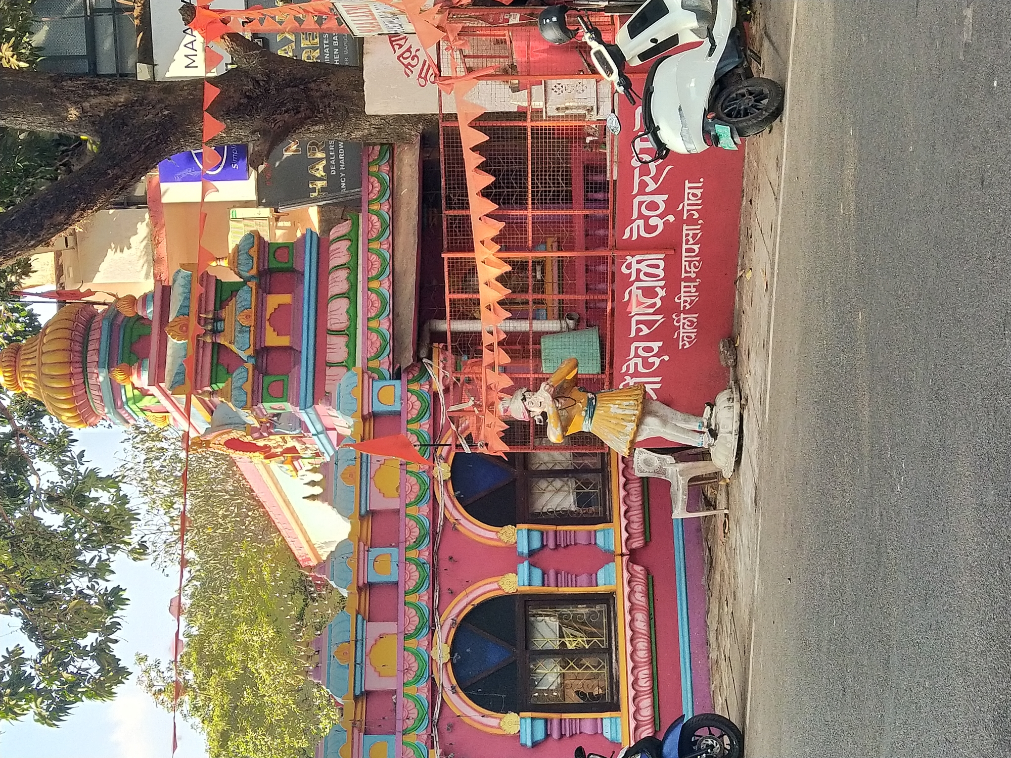 The image shows a colorful Hindu temple in Mapusa, Goa, India. The temple has a brightly painted facade with intricate carvings and decorations. Orange pennants are strung across the front of the temple. A white electric scooter is parked in front. A life-sized statue of a person dressed in traditional Indian clothing is placed next to the temple entrance. A sign on the wall reads "श्रीदेव राष्ट्रोळी देवस्थान, खोर्ली सीम, म्हापसा, गोवा" which translates to "Shree Dev Rashtroli Devasthan, Khorli Seem, Mapusa, Goa." The temple is situated next to a road. Trees are visible on the left side of the image. Another sign is visible on top of the temple.