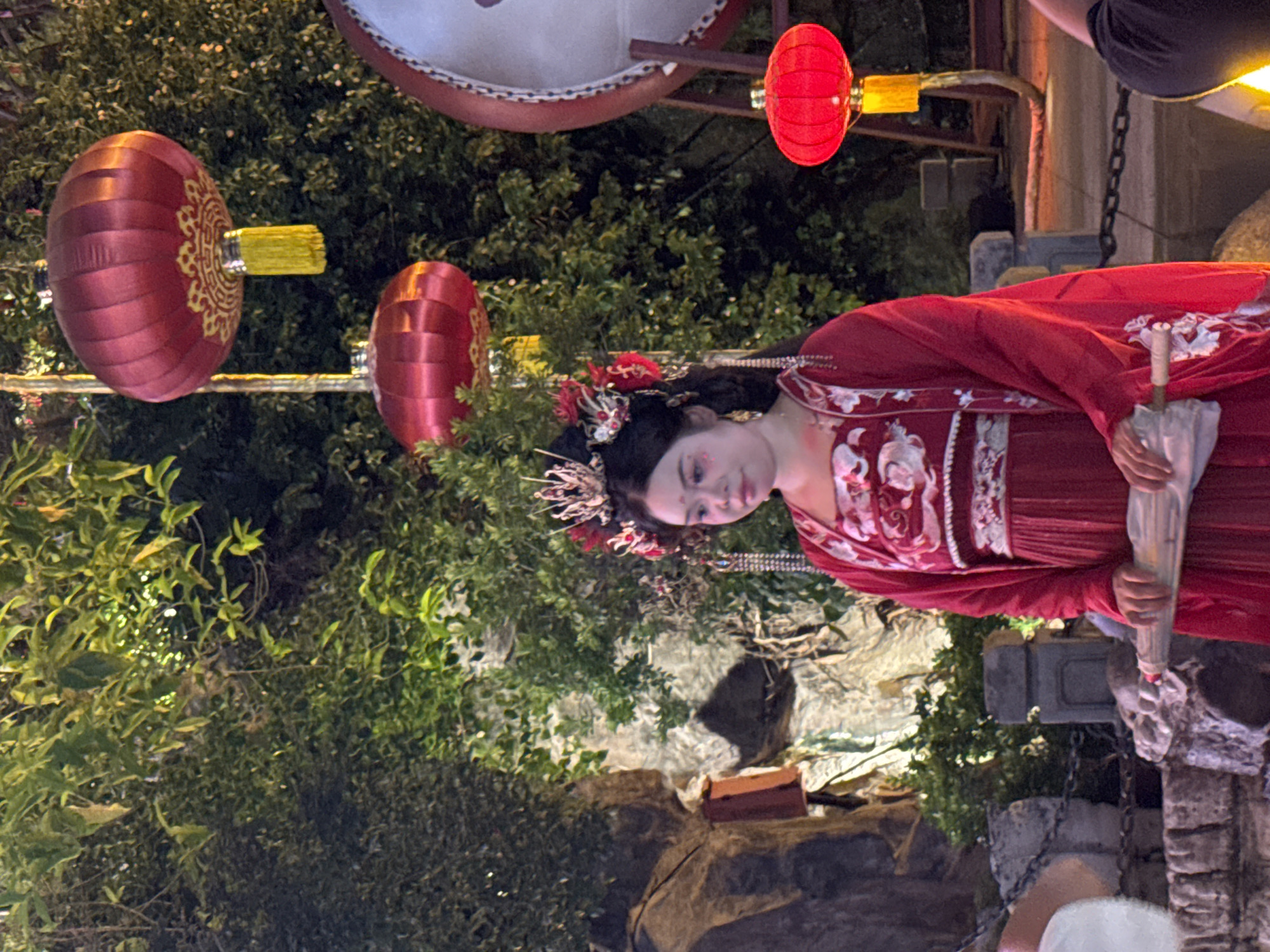 A woman in traditional red Chinese clothing with an elaborate headdress stands on a stone pathway. She is holding a closed white umbrella and appears to be looking down. The background features lush green foliage, a large red lantern hanging from a pole, and a section of a large drum. The scene is illuminated, creating a dramatic and festive atmosphere. The woman is wearing heavy makeup, with red accents on her cheeks and eyes. The clothing is richly embroidered with white floral patterns. The setting suggests a cultural performance or celebration.