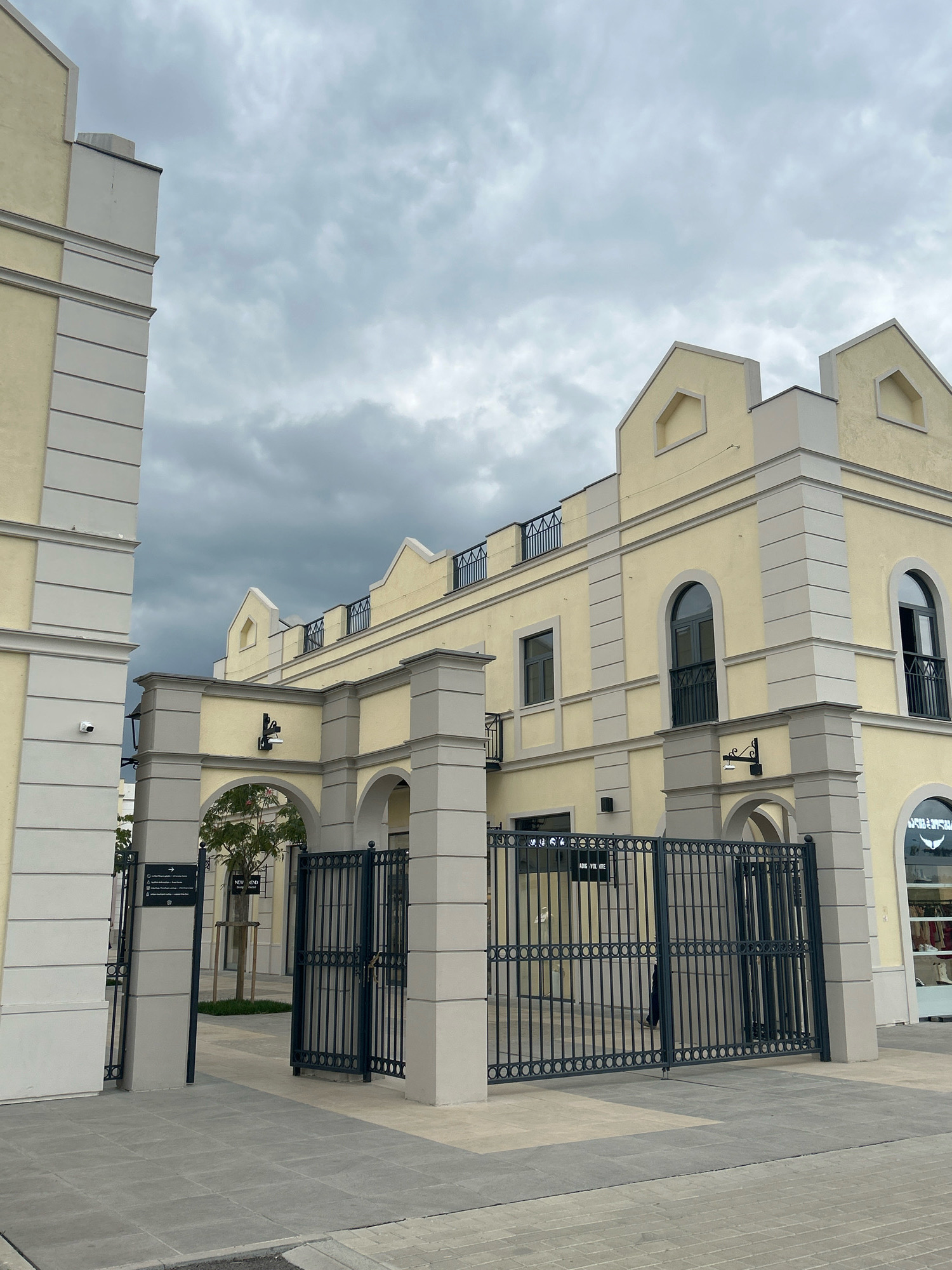 The image shows the exterior of a building with a pale yellow facade, featuring architectural details in a lighter beige. The building has a distinctive roofline with crenellations and arched windows, some of which have black wrought-iron balconies. A tall, dark metal gate with an ornate pattern is open, leading into a courtyard. Two stone pillars flank the gate. To the left of the gate, a signpost with directional arrows and text is mounted on a similar stone pillar. A security camera is visible on the left side of the building. The ground is paved with grey rectangular tiles. Beyond the gate, a small tree with green foliage is visible on a patch of grass, with arched openings of the building visible in the background. The sky is overcast and cloudy. The location is identified as Gamarjveba, Georgia.