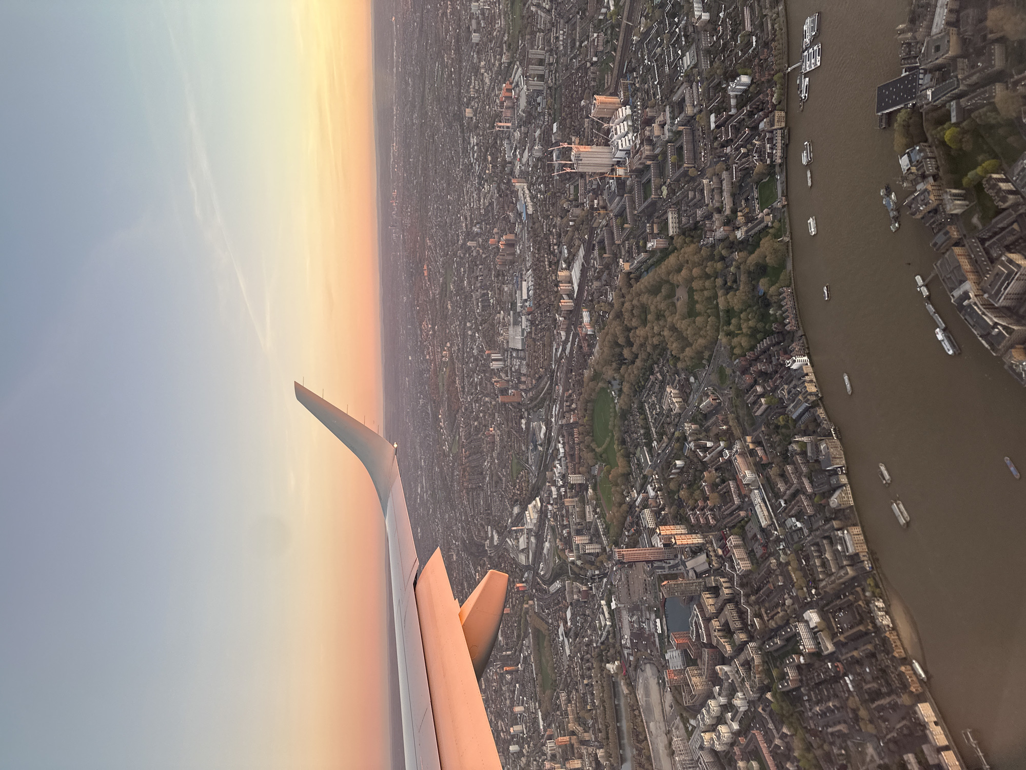 Aerial view from a plane, showing part of London, United Kingdom. The wing of the plane is in the foreground. Below is a dense urban landscape, bisected by the River Thames. Numerous buildings, streets, and green spaces (parks or gardens) are visible. The river contains various vessels, including boats and barges. The sky is tinted with the colors of sunrise or sunset.