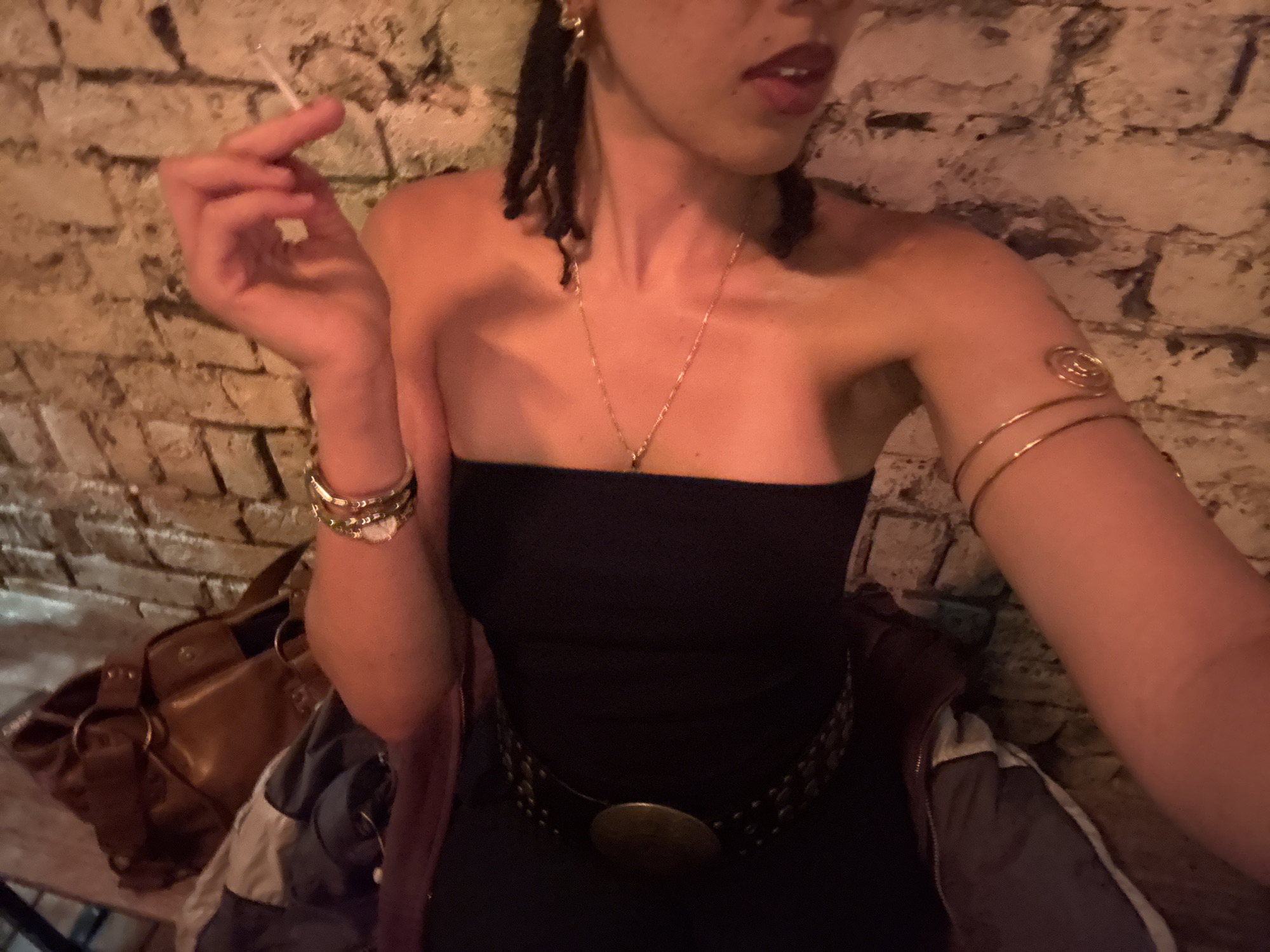 A medium close-up shot captures a person leaning against a textured, light-colored brick or stone wall in dim, warm lighting, suggesting a nighttime setting. The subject, likely female, has dark dreadlocks visible on the left side of their head and is wearing dark red lipstick. They hold a lit cigarette between the thumb and forefinger of their left hand.
The person is dressed in a black strapless top. Several gold accessories adorn them, including delicate gold earrings, a thin gold chain necklace with a small pendant, multiple gold bangles on their left wrist, and a spiral-designed gold arm cuff on their right upper arm. A black belt with a large, circular, bronze-colored buckle and stud details is worn around their waist.
To their left, a brown leather shoulder bag or purse is visible, and beneath them rests a jacket or coat with sections of grey, white, and dark blue fabric. The overall atmosphere is casual and relaxed. No text is visible in the image. The provided context indicates the location is London, United Kingdom.
