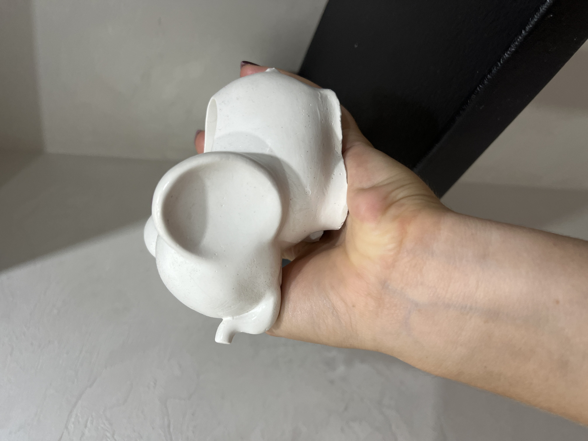 A person's hand is holding a white, elephant-shaped ceramic pot against a light gray textured wall and a dark shelf. The pot has large ears and a hollow interior, suggesting it is a planter or decorative item. The hand is positioned to support the pot from underneath and the side. The background appears to be indoors, possibly a home or studio setting.
