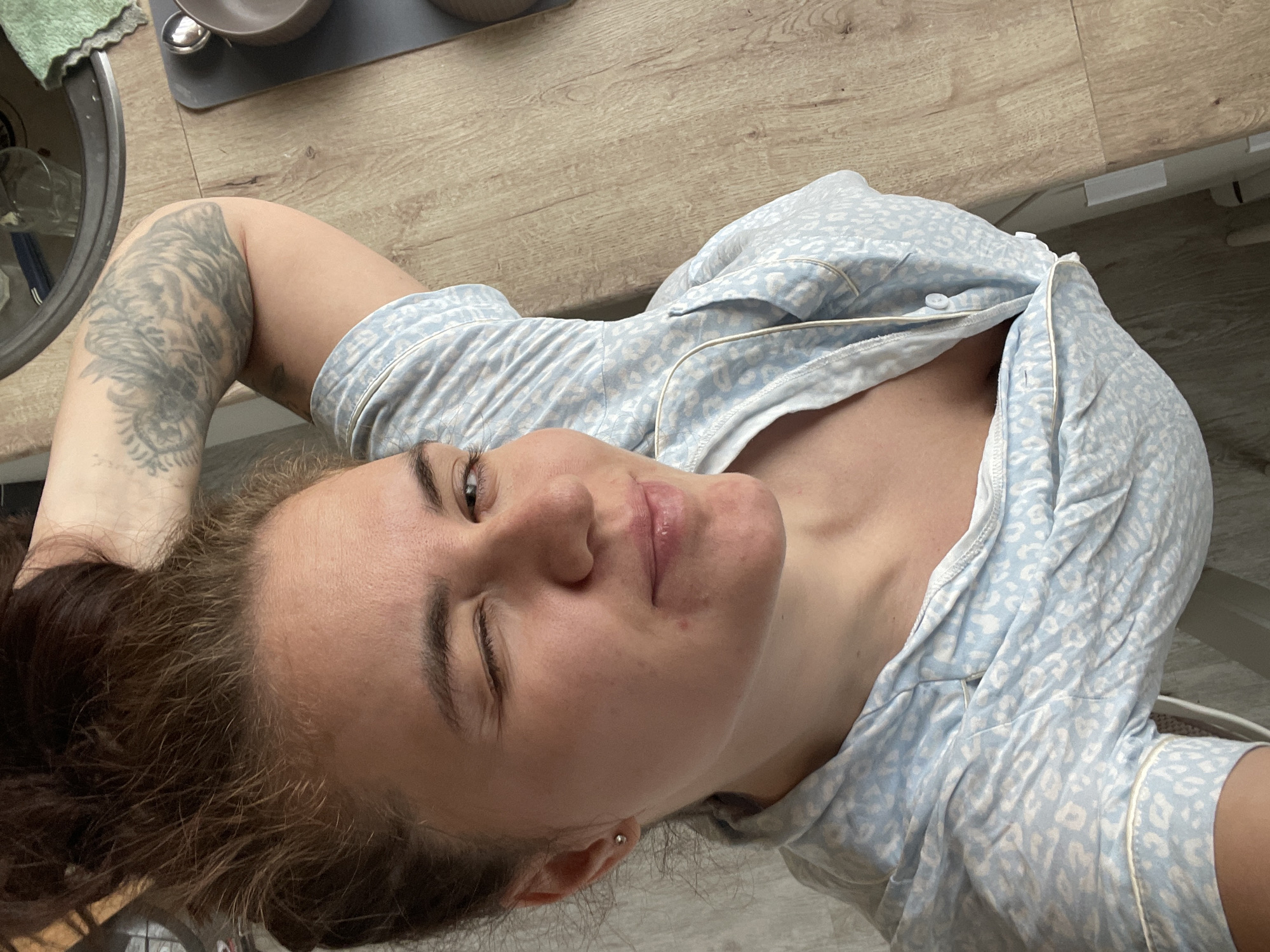 A woman with brown hair and a tattoo on her left arm is lying on a surface with a light brown wood-grain pattern. She is wearing a blue and white patterned pajama top. Her eyes are partially closed, giving the appearance of a wink. A stainless steel sink and bowls are visible in the upper left corner. The background includes a light grey surface. There is no evidence to support the presence of the location in the city of Мосметаллоконструкция Онт, Russia.