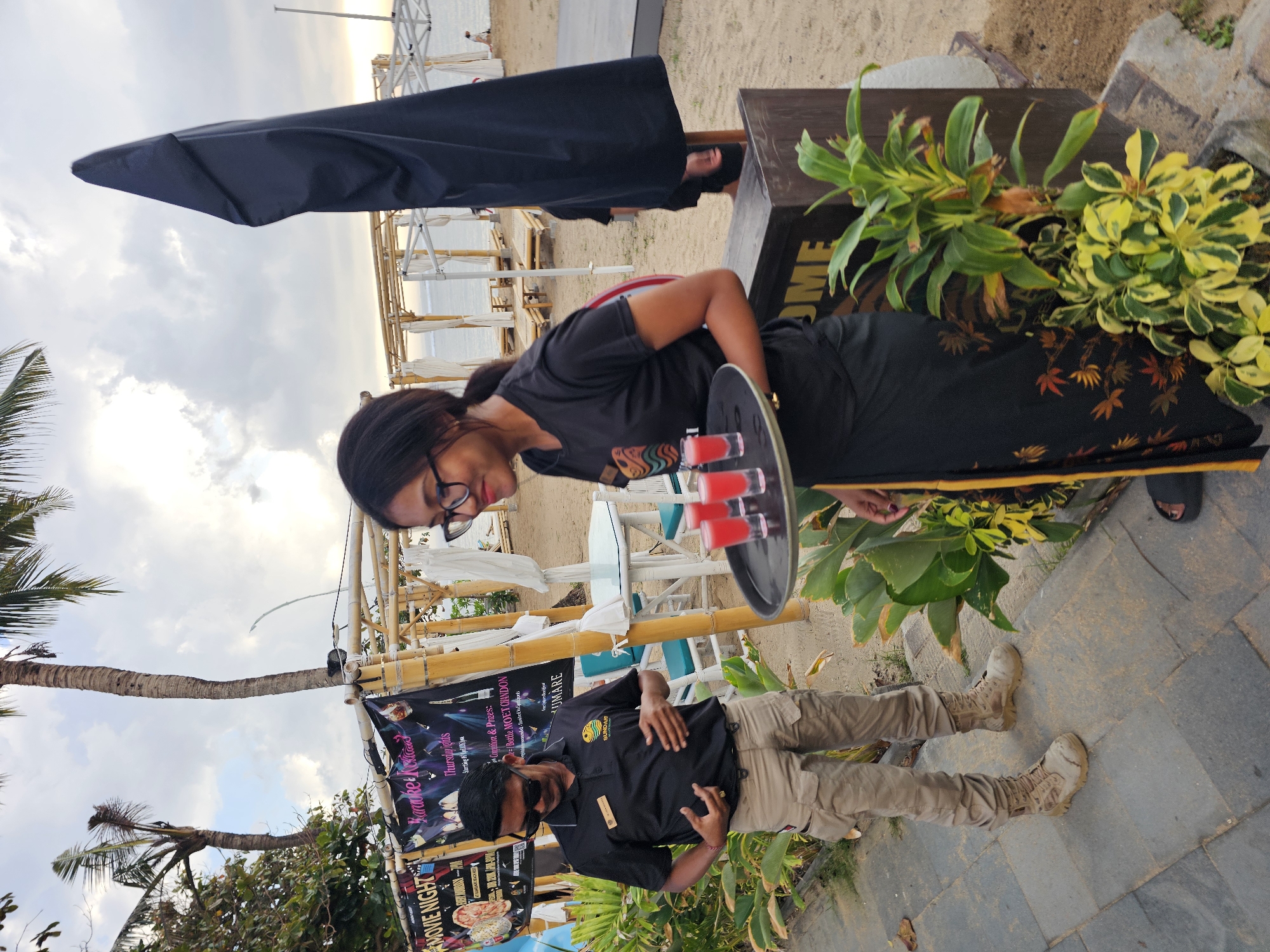 A young woman, wearing glasses and a black t-shirt with a patterned skirt, stands on a sandy beach pathway in Denpasar, Indonesia. She is holding a tray with four small red drinks. To her left, a man in sunglasses, a black polo shirt with a logo, and khaki cargo pants is looking down at his hands. Behind them, palm trees, bamboo structures, white tents, and beach chairs are visible, suggesting a resort or entertainment venue. The sky is overcast.