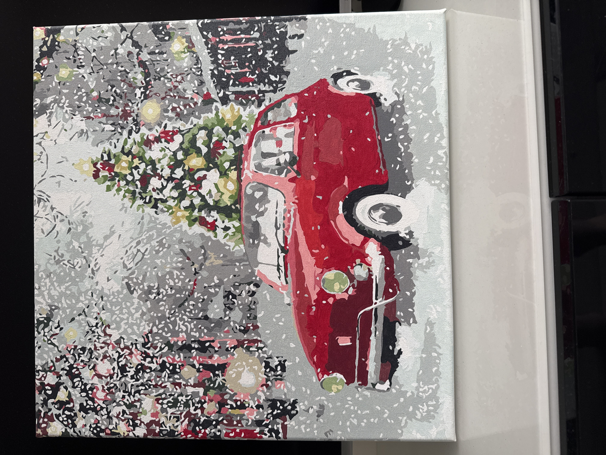 The image depicts a painting on canvas of a red vintage car parked in a snowy environment. The car is the central subject, with a Christmas tree decorated with lights and ornaments visible behind it. The background suggests a festive winter scene with trees adorned with lights and falling snow. The style of the painting is impressionistic, with bold strokes and a somewhat simplified representation of details. The painting is framed and displayed on a reflective white surface, with a black background behind it. There are no people or animals visible in the painting. The scene is a generic winter holiday depiction and does not provide any specific landmarks or context to suggest it is located in Tallinn, Estonia.