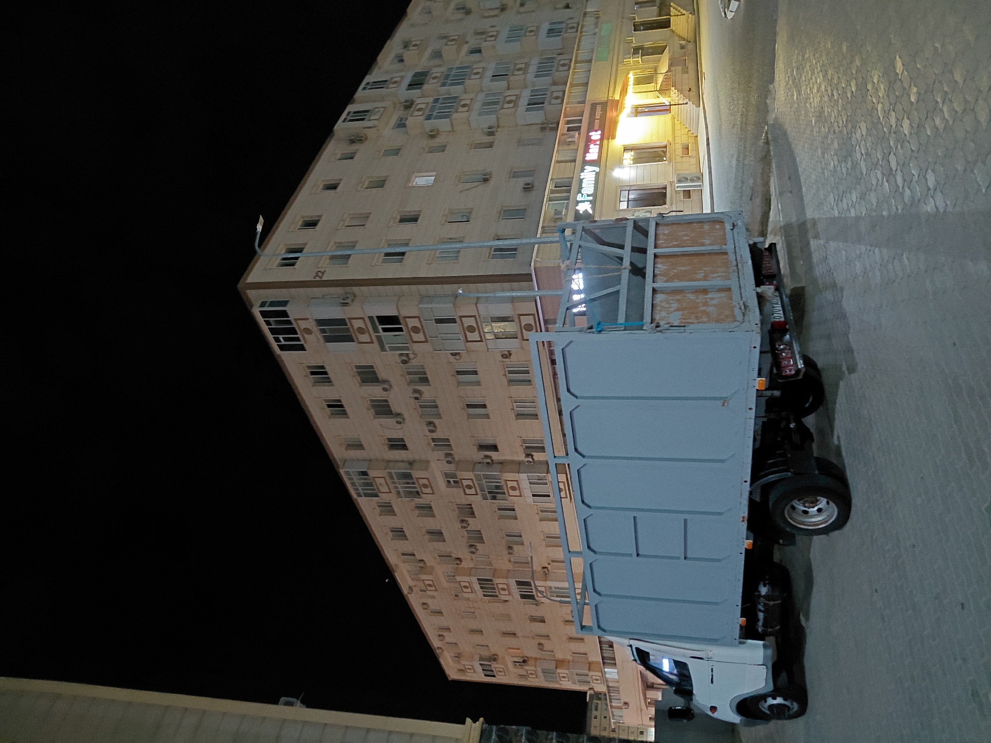 An outdoor, nighttime, low-angle shot captures a light-colored delivery truck parked on a cobblestone street in front of a large, multi-story apartment building. The building, possibly located in Aktau, Kazakhstan, features numerous windows and air conditioning units. A neon sign with the words "Family Market" is visible on the ground floor of the building. The truck has a light blue cargo box and appears to be empty. The street is illuminated by streetlights and the lights from the building, casting shadows on the pavement.