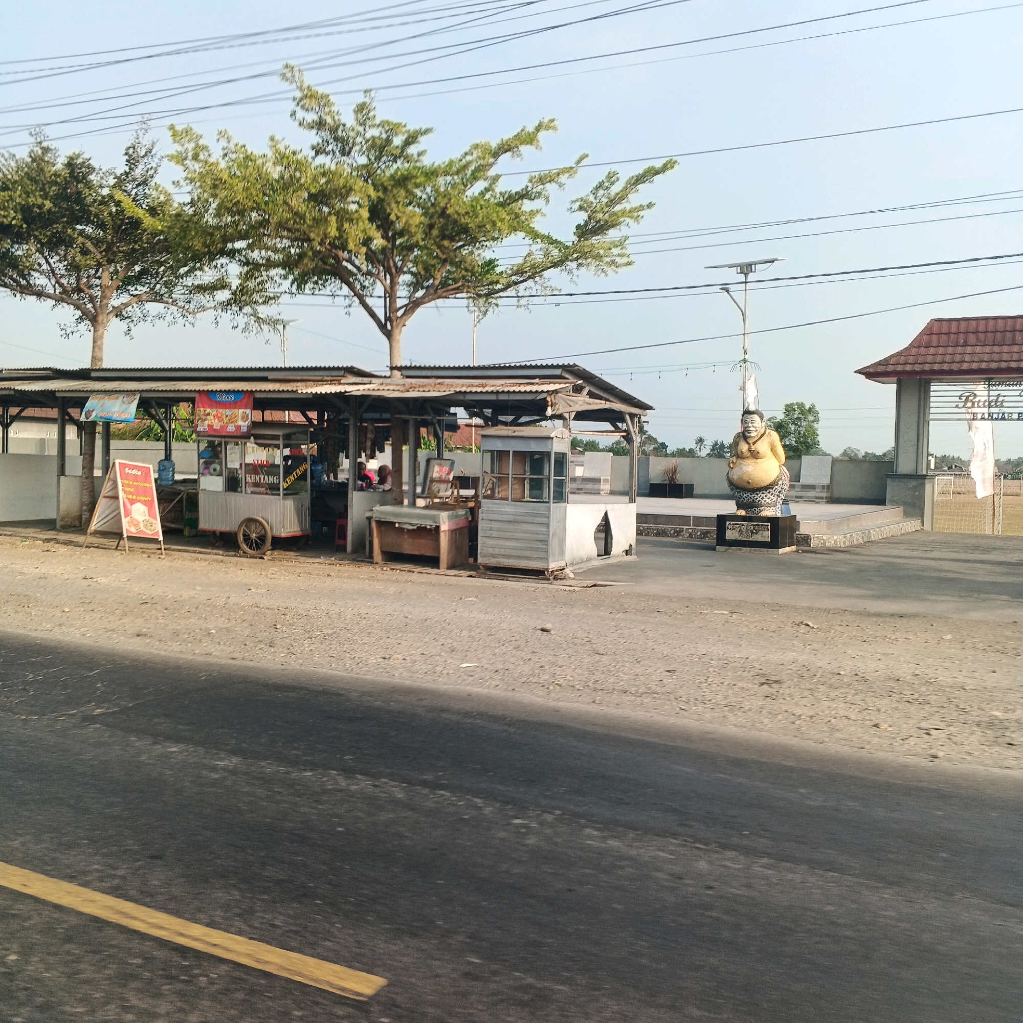 The image shows a roadside scene in Menganti, Indonesia. In the foreground, a paved road with a yellow line is visible. Behind the road, there is a dirt area and a small food stall with a corrugated metal roof. The stall has several carts, signage, and what appears to be bottled water. A white A-frame sign with text and images is placed in front of the stall. Further in the background, there is a statue of a rotund figure on a pedestal, and beyond that, an entrance gate with the words "Taman Budaya Banjarejo" visible. Trees with sparse green foliage are present throughout the scene, and electrical wires are strung across the sky. The sky is clear and light blue, suggesting it is daytime. There are a few people visible in the background near the food stall, but their identities are not clear. The overall scene depicts a typical street-side commercial area.