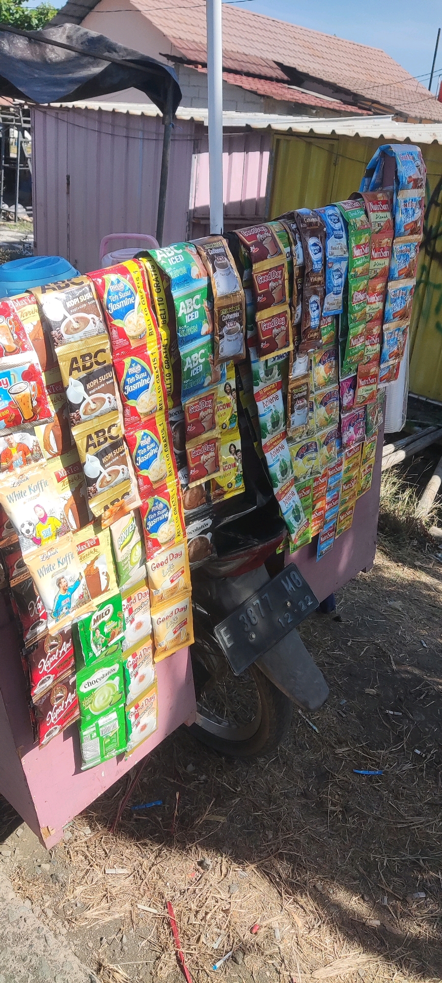 The image depicts a roadside stall selling various packaged beverages in Kalimaro, Indonesia. The stall is mounted on a motorcycle, evident from the visible rear tire, fender, and license plate which reads "E 3877 MO 12 22". The stall is laden with rows of individually packaged instant drinks, including coffee, tea, and chocolate flavored beverages. Brands like "ABC," "Good Day," "NutriSari," "Milo," and "White Koffie" are clearly visible. The stall structure itself appears to be a pink-colored cabinet or box, partially covered by a dark tarp. In the background, a pink building with a corrugated metal roof is visible, along with some vegetation. The ground is covered with dry grass and debris. No people are present in the immediate vicinity of the stall. The lighting suggests it is daytime, with clear shadows cast on the ground.