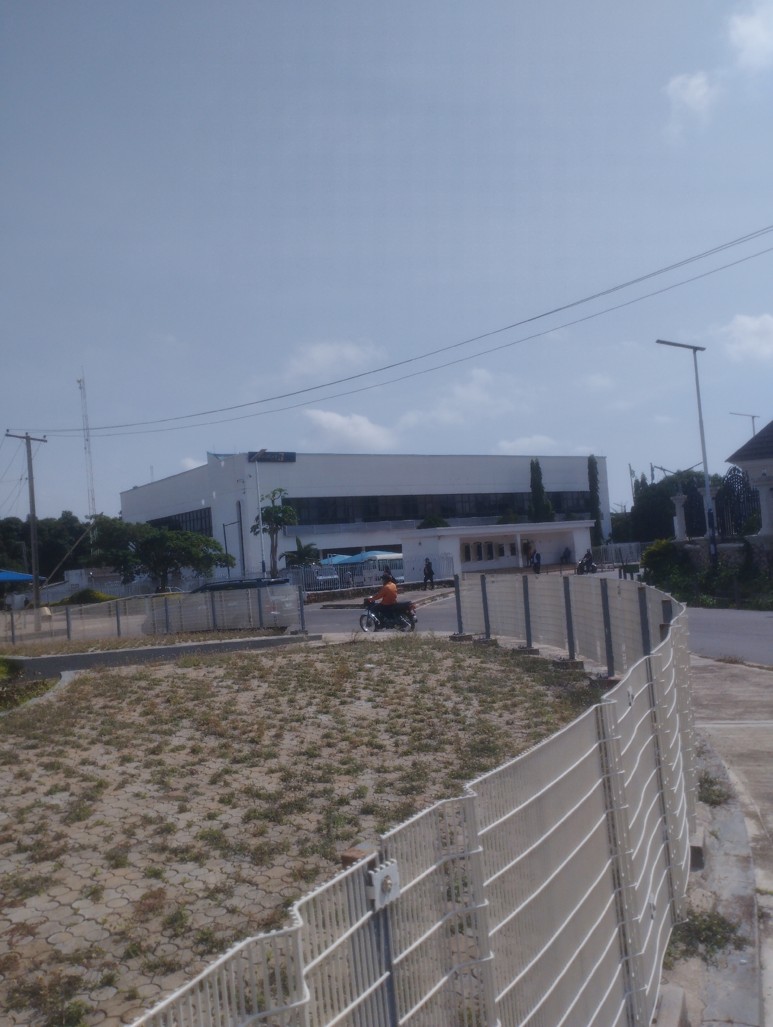 The image depicts a wide view of a street scene in Jos, Nigeria, dominated by a modern, multi-story white building with dark windows. A white metal fence runs along the foreground, separating a dry, sparsely vegetated area from a paved walkway. On the walkway, a person is riding a motorcycle. The sky is pale blue with scattered clouds. In the background, trees and a tall antenna are visible. The overall impression is one of an urban or semi-urban environment under daylight conditions.