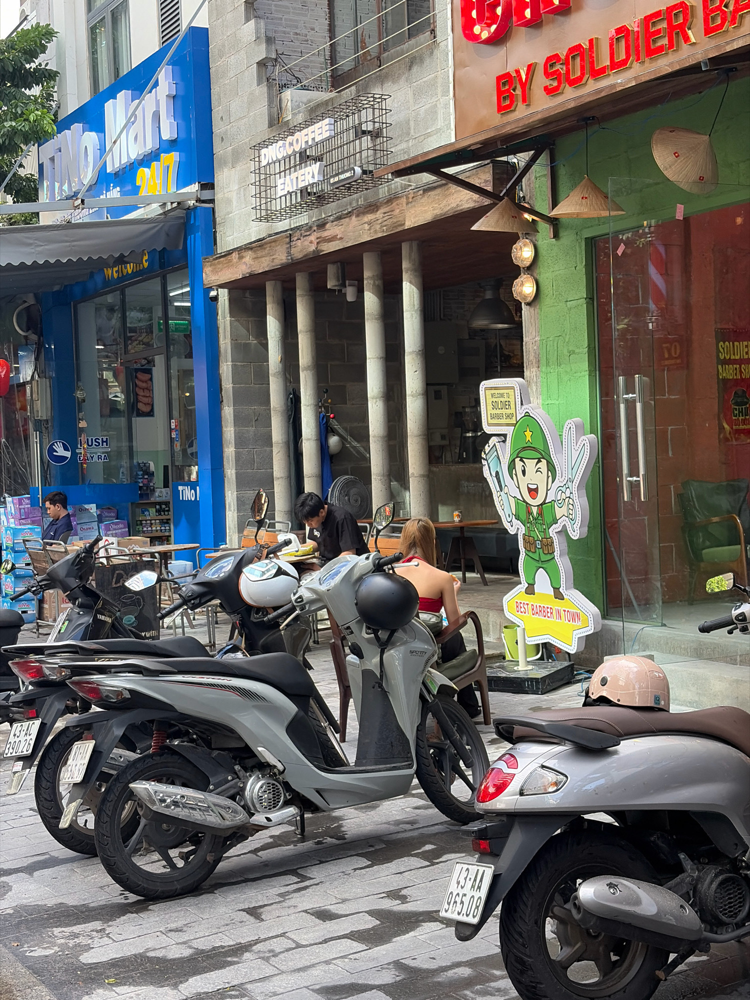In the city of Da Nang, Vietnam, on a street, a barber shop named "BY SOLDIER" is prominently displayed with red lettering. In front of the shop, a cartoon mascot of a barber holding scissors and a clipper stands on a sign that reads "BEST BARBER IN TOWN." Adjacent to the barber shop is a store labeled "Tino Mart 24/7," with a blue facade. Several motorbikes are parked on the paved street, with a light gray scooter in the foreground displaying a license plate. A person with blond hair wearing a red top is seated on one of the parked scooters, facing away from the camera. Inside Tino Mart, two men are visible, one sitting and looking at a laptop, the other standing. The street is lined with buildings and various shop signs.