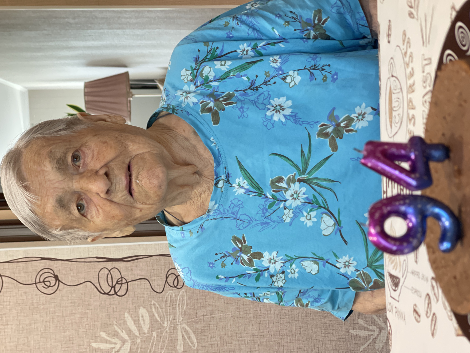 An elderly woman with gray hair is shown from the chest up, looking directly at the camera. She is wearing a bright blue shirt with a floral pattern of white and green flowers and brown accents. Two candles, shaped like the numbers "9" and "4" in a gradient of purple to blue, are placed on a chocolate cake in front of her. The cake is on a tablecloth that has a coffee-themed pattern, including drawings of coffee beans and a cup. The background shows a wall with a textured wallpaper, a lamp with a pink shade on a stand, and a curtain with a brown swirled pattern. The scene appears to be indoors, likely in a home setting.